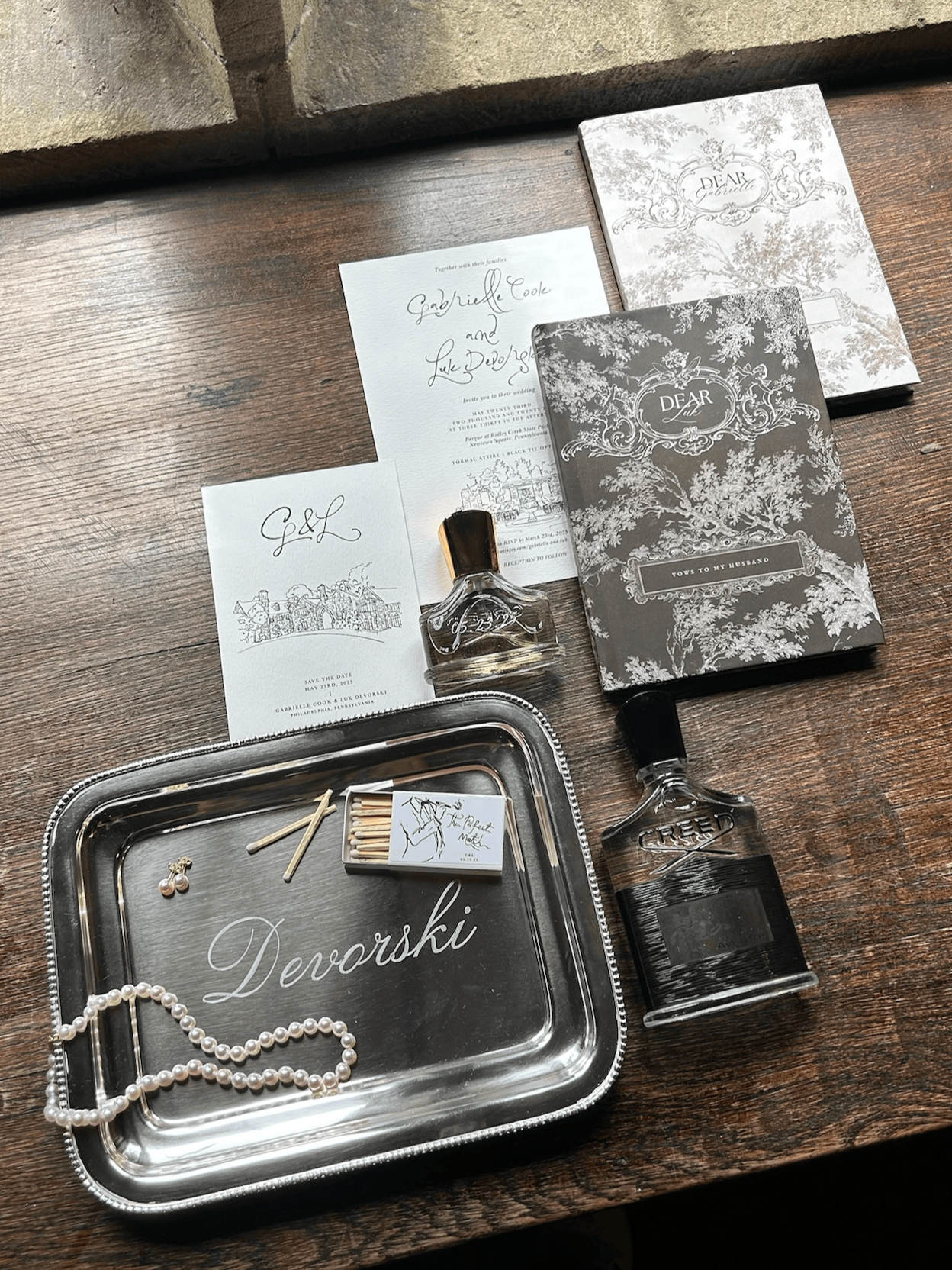 A collection of wedding items on a wooden surface, including a silver tray with pearl necklace, matches, and earrings, wedding invitations, vow booklet, and perfume bottles.