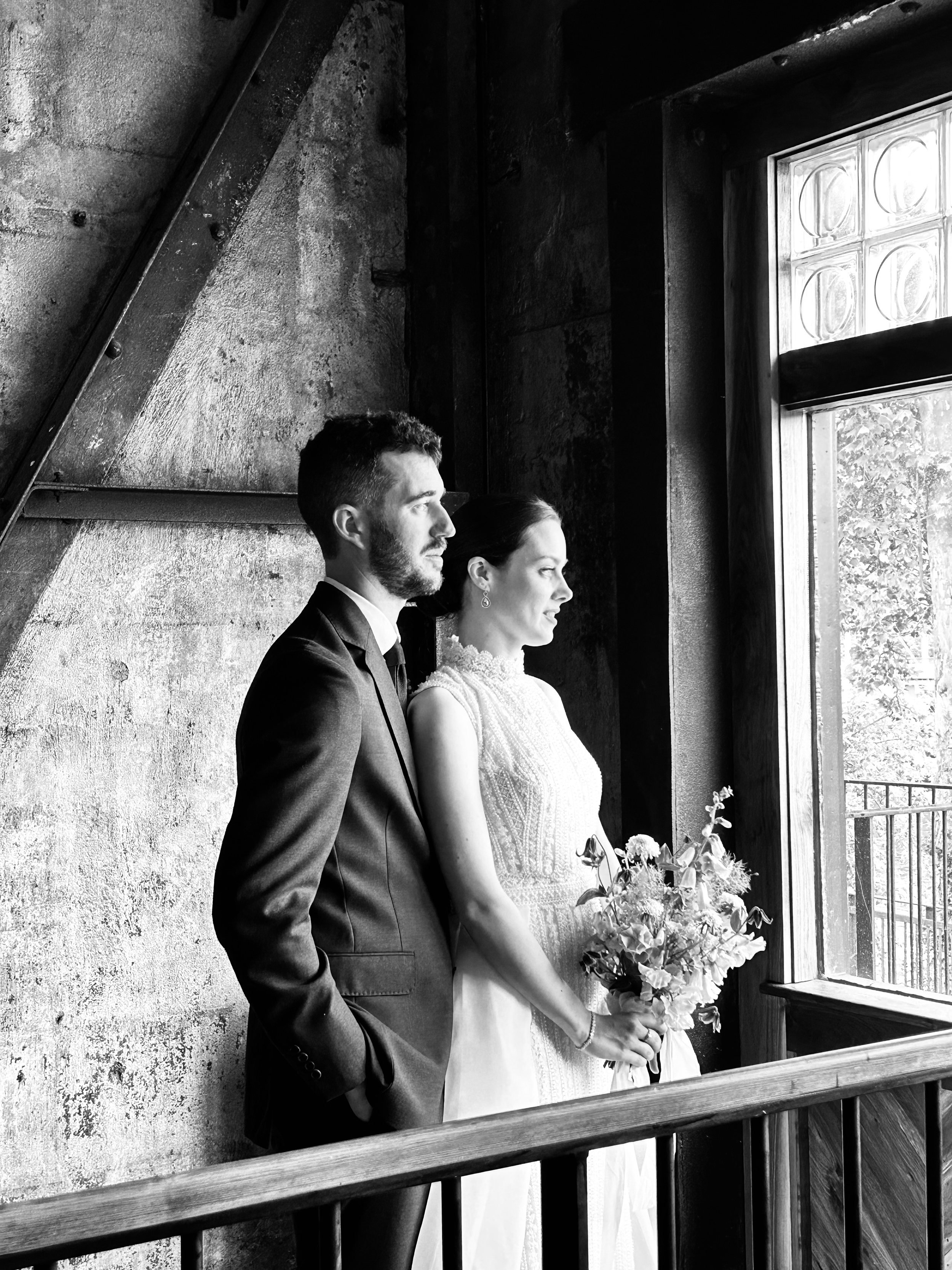 A black and white photo of a bride and groom standing close together, gazing out a window. The bride holds a bouquet, and both are dressed in formal wedding attire.