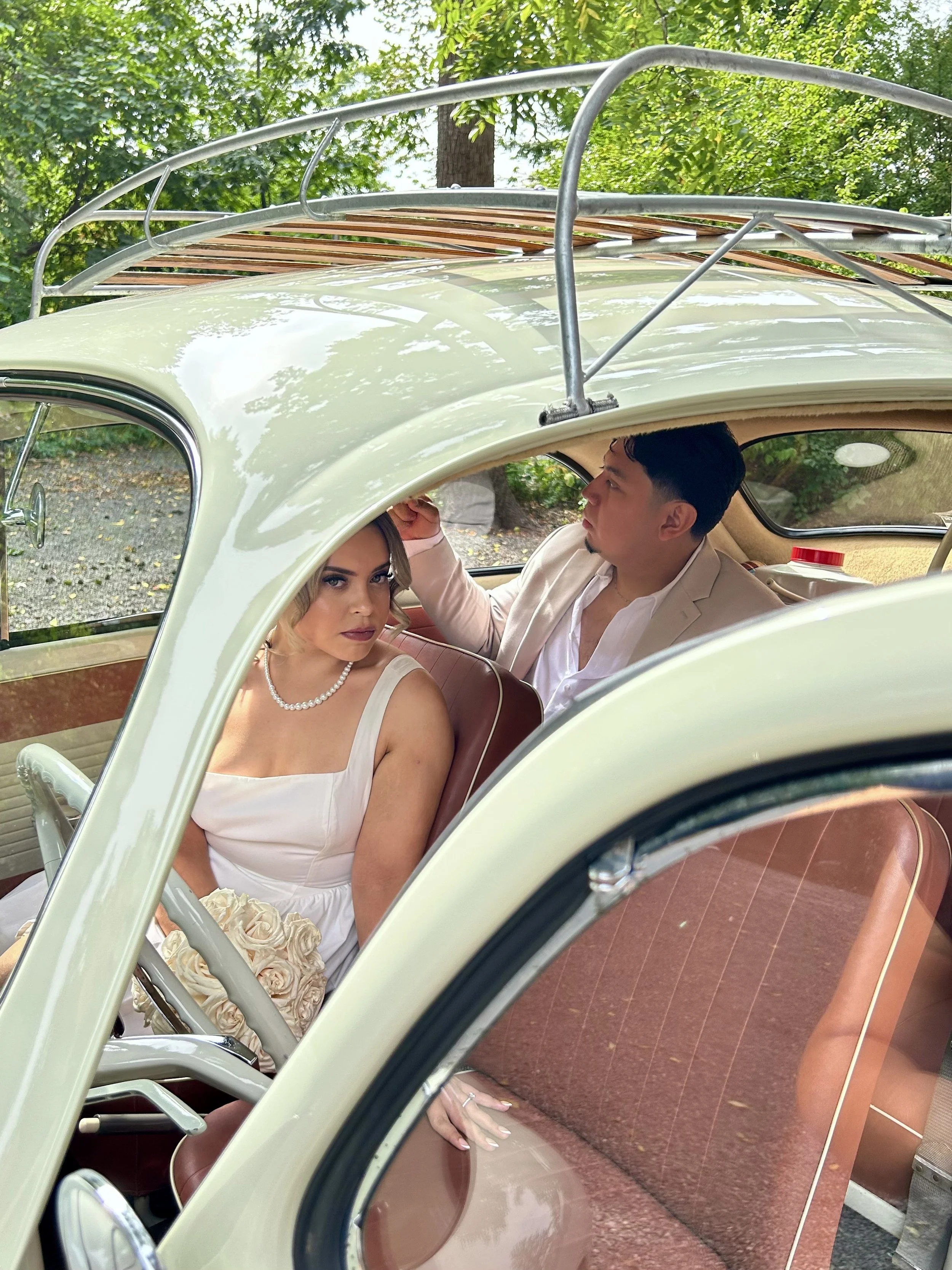 A woman in a white dress with a pearl necklace sitting inside a vintage white car with tan interior, holding a bouquet of cream roses. A man in a light-colored suit is sitting beside her, looking at her and touching her hair, with green trees outside the car window.