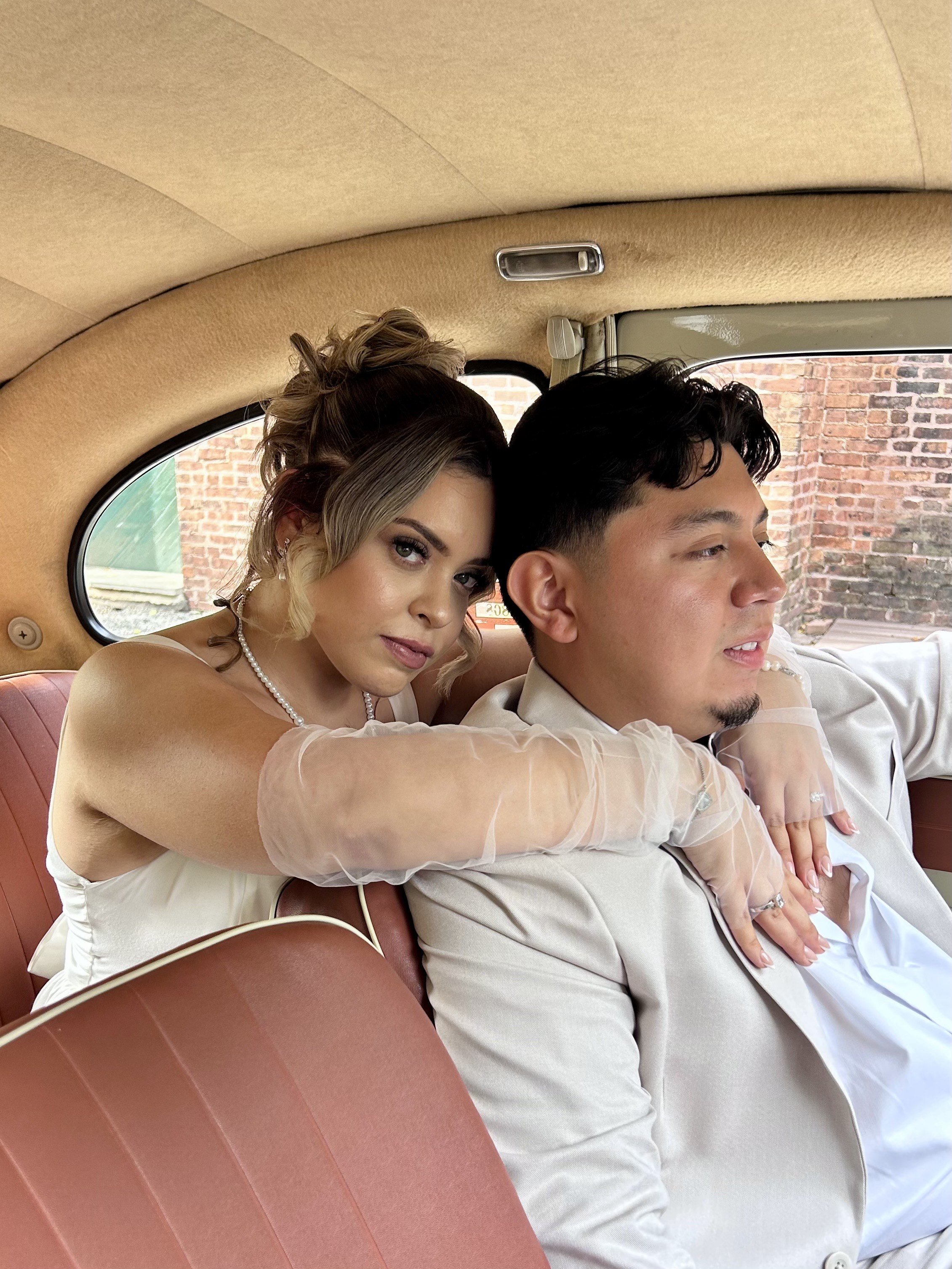 A woman and a man sitting in the back of a vintage car. The woman has blonde, curly hair styled in an updo, wearing a white dress, pearl necklace, and sheer gloves. The man has short dark hair, a slight beard, and is dressed in a light-colored suit with a white shirt. The woman is leaning on the man's shoulder with her arms around his neck, both looking relaxed. The background visible through the car window shows brick walls.