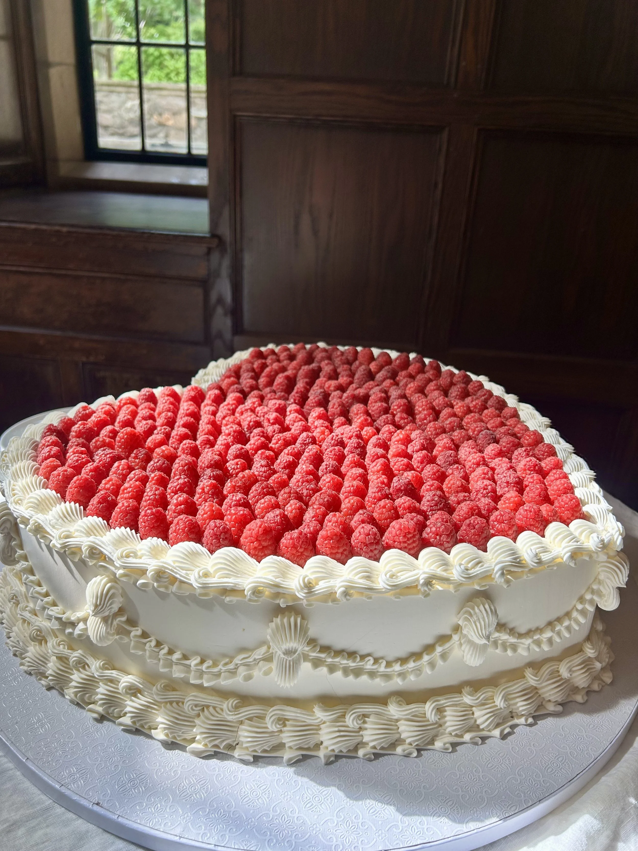 A large heart-shaped cake decorated with fresh raspberries on top and white frosting borders around the edges, placed on a white decorative cake board.