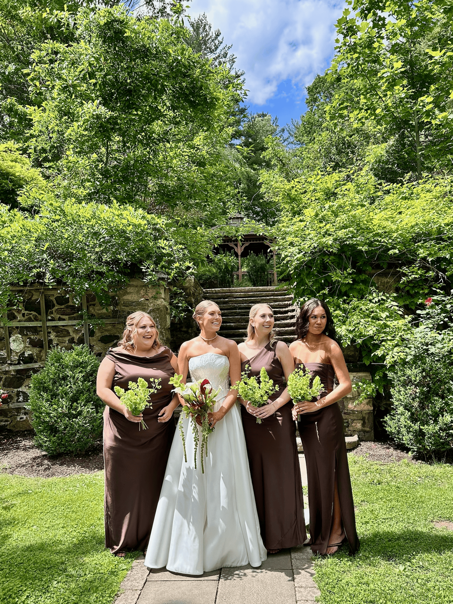 A bride in a white strapless wedding gown holding a bouquet standing next to four bridesmaids in dark brown dresses, all holding small bouquets, in a lush green garden with stone steps behind them.