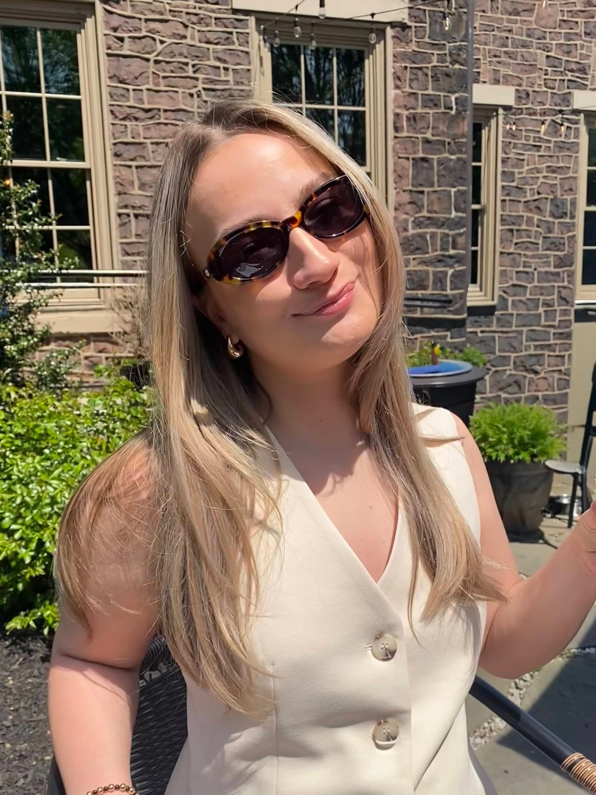 A woman with blonde hair wearing sunglasses and a sleeveless beige top with large buttons, smiling outdoors in front of a brick building with windows and plants.
