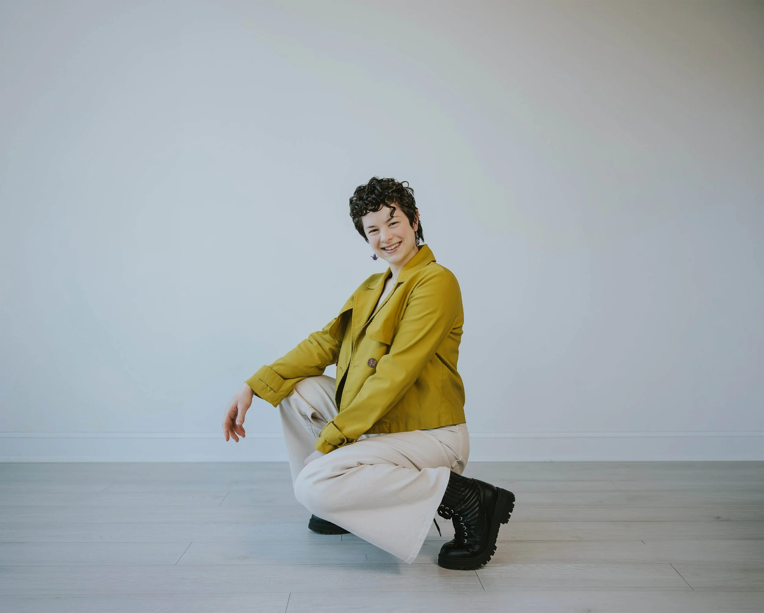 A person with short, curly dark hair and a bright smile, wearing a yellow jacket, beige pants, and black combat boots, is crouching on one knee on a light wooden floor against a plain white wall.