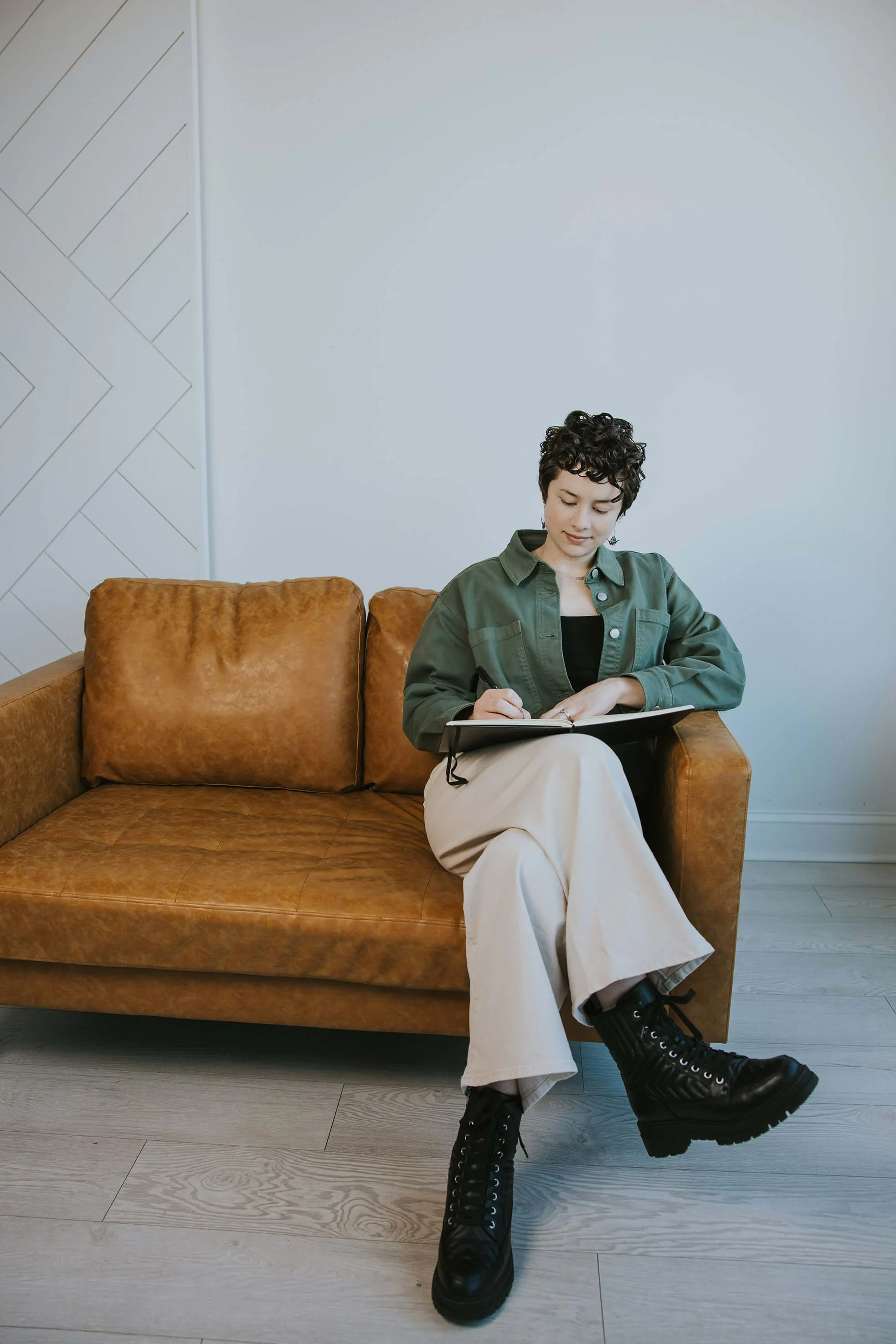 A young woman with short curly hair sitting on a brown leather sofa, writing in a notebook with a pen in a room with light-colored walls and light wood flooring, wearing a green jacket, black top, beige wide-leg pants, and black combat boots.