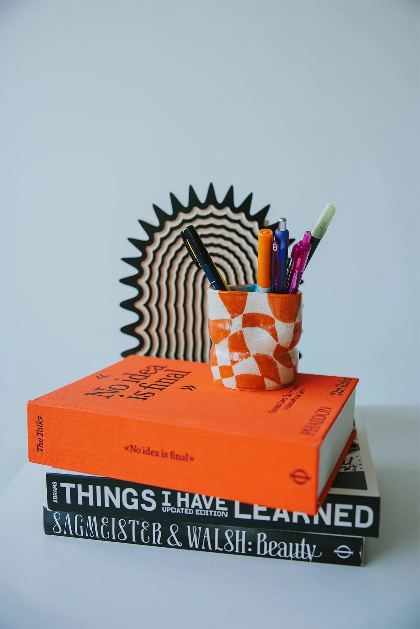 A stack of three books with a small, orange and white patterned cup holding colorful pens resting on top. Behind the cup, there is a black and white patterned decorative object, all set against a plain light background.