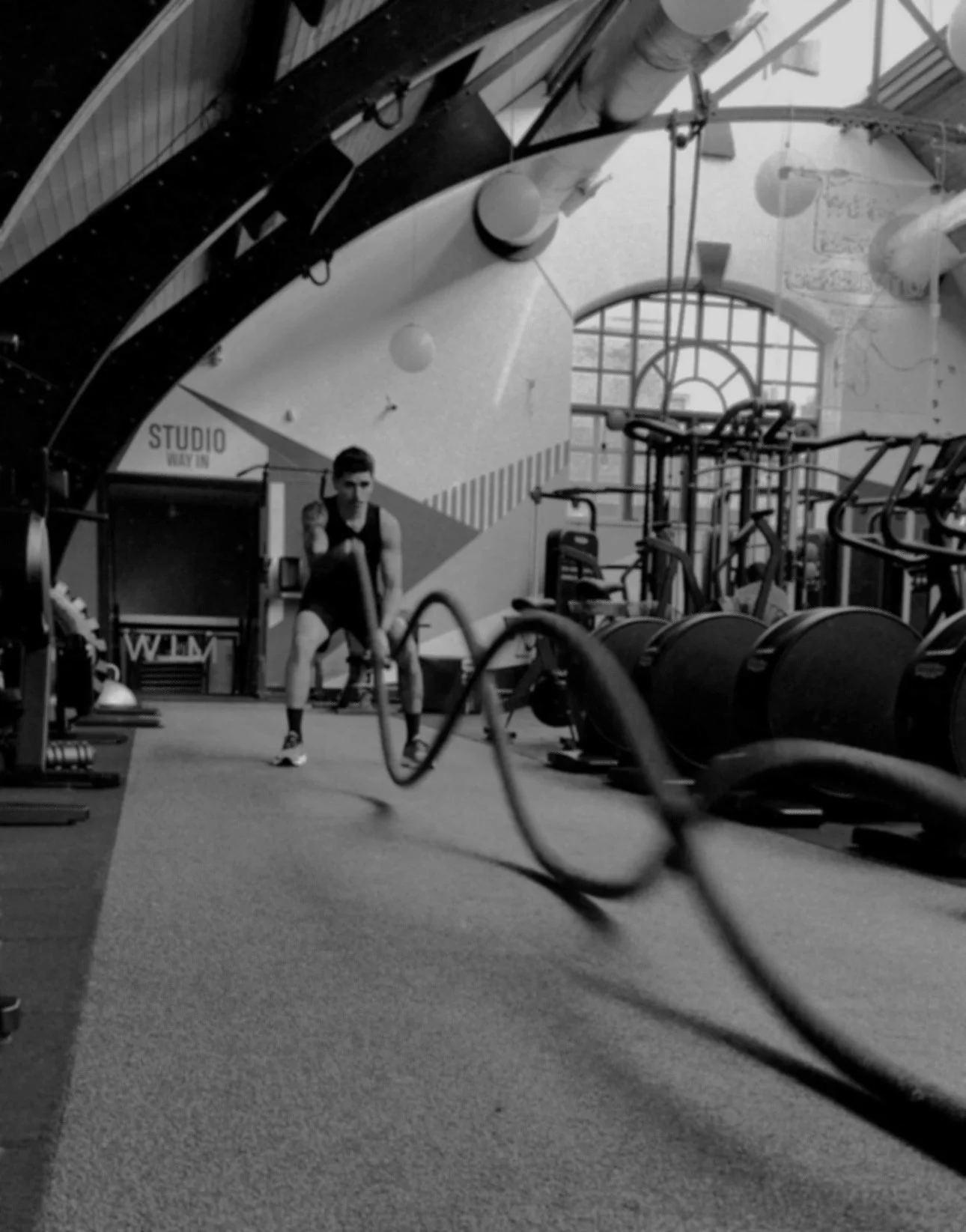 Exercises with battle ropes in a gym, black and white image with workout equipment and a large window in the background.