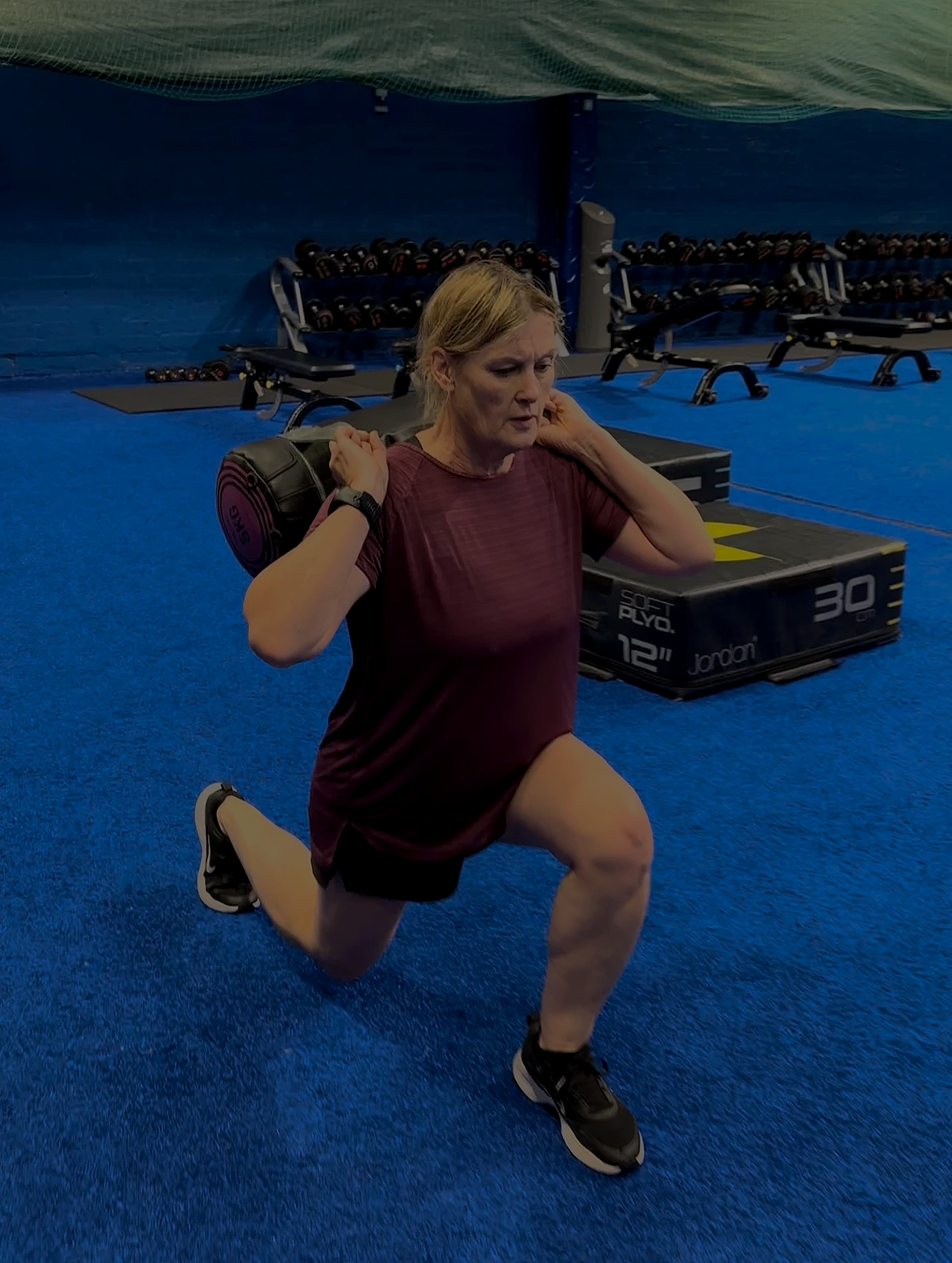Woman performing a lunge exercise while holding a weighted barbell on her shoulders in a gym with blue carpet and workout equipment in the background.