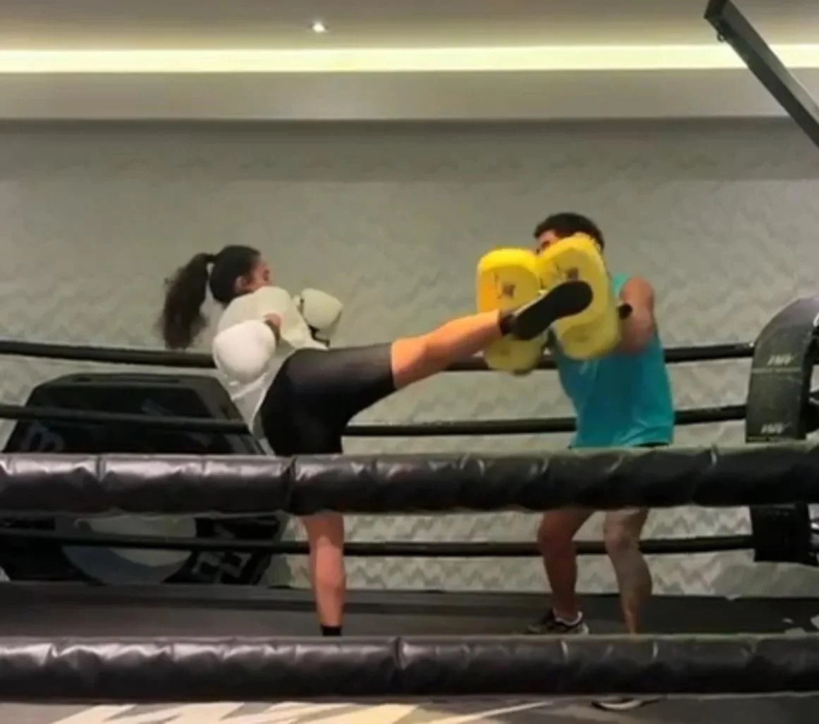 A woman practicing boxing in a ring, delivering a high kick to a man holding yellow punching mitts, with both focused on the training exercise.