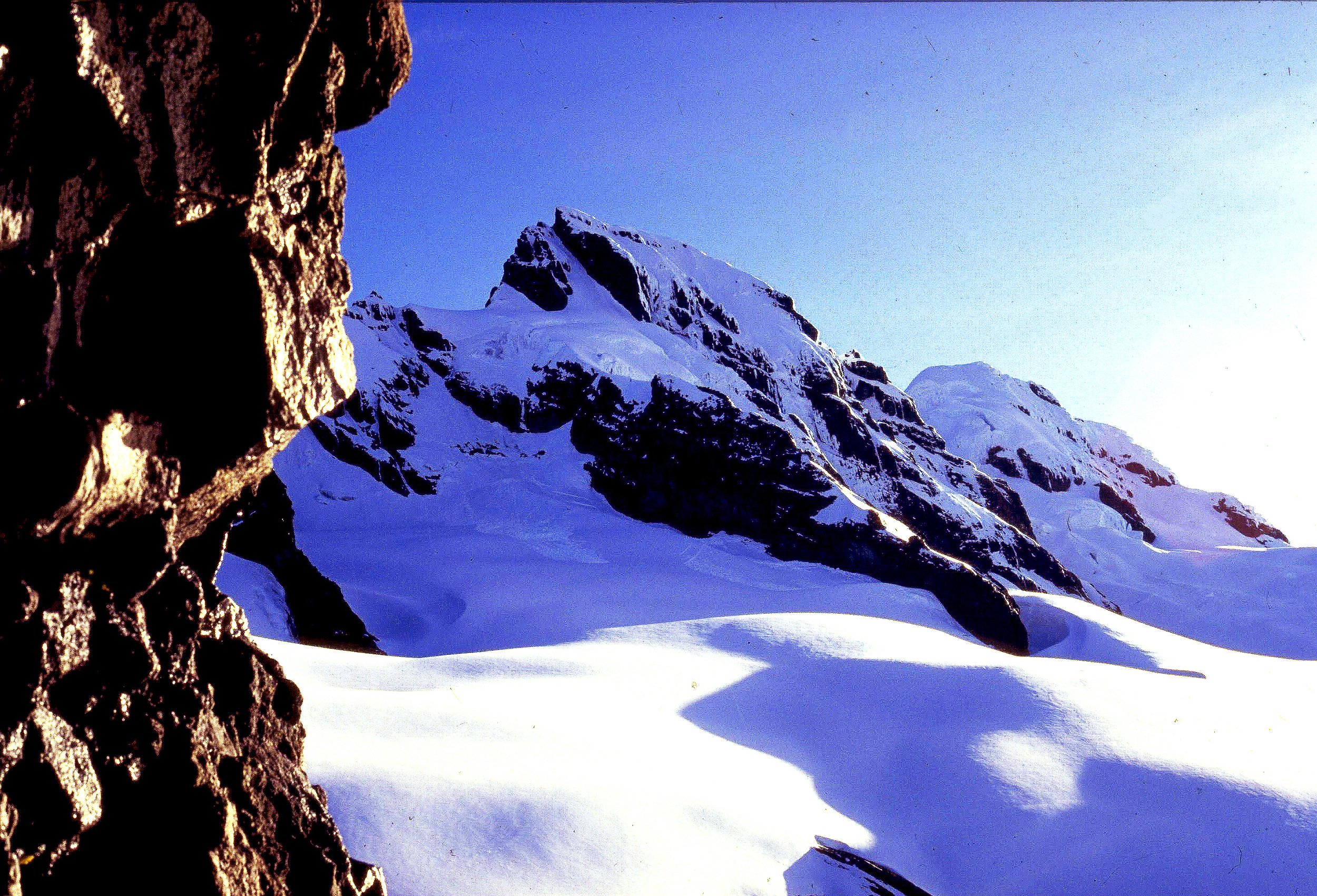 Snow-covered mountain peaks with a clear blue sky, seen from a rocky foreground.