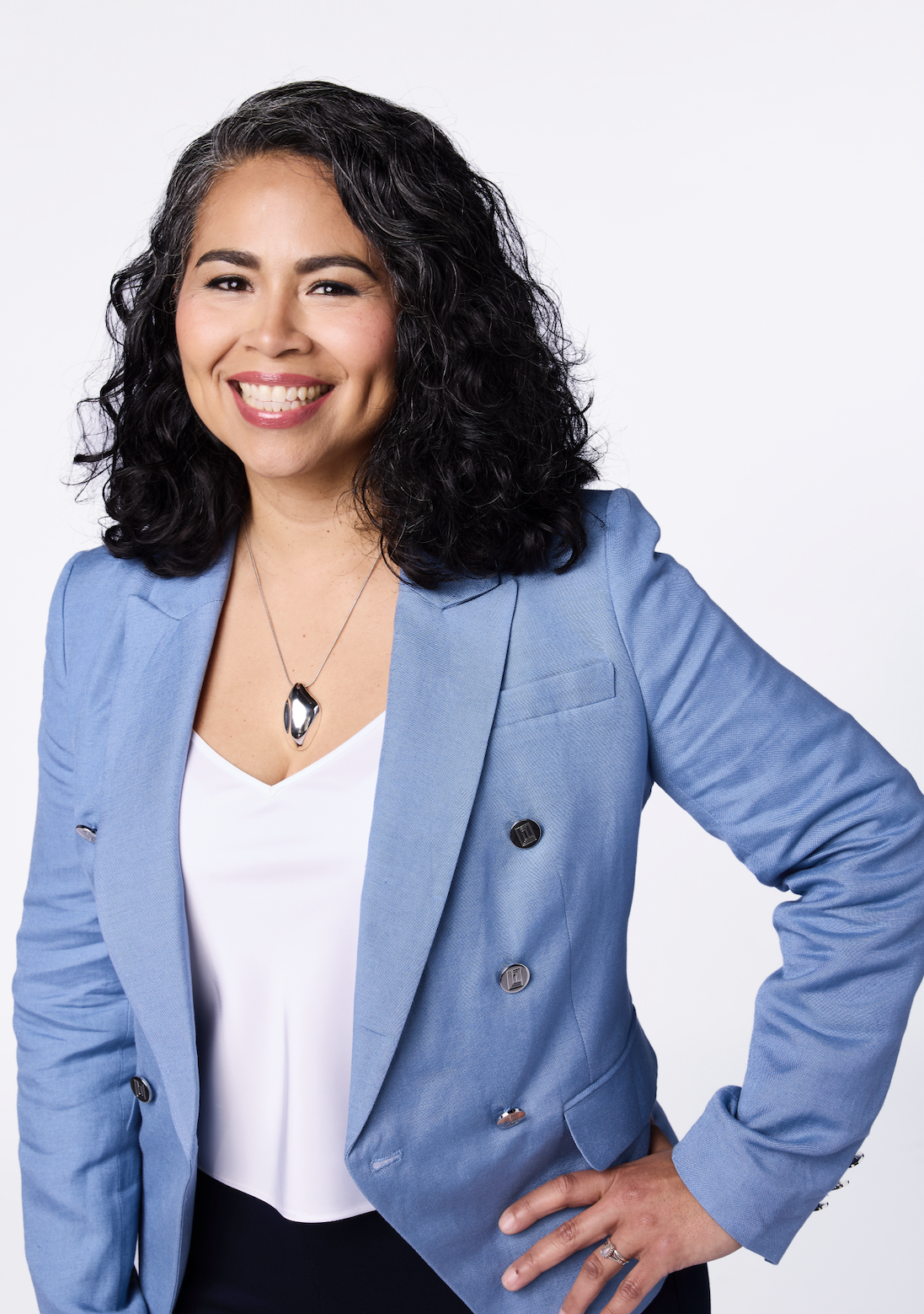 A woman with dark curly hair wearing a light blue blazer, white top, black pants, and a silver necklace smiling confidently, posing with one hand on her hip against a white background.