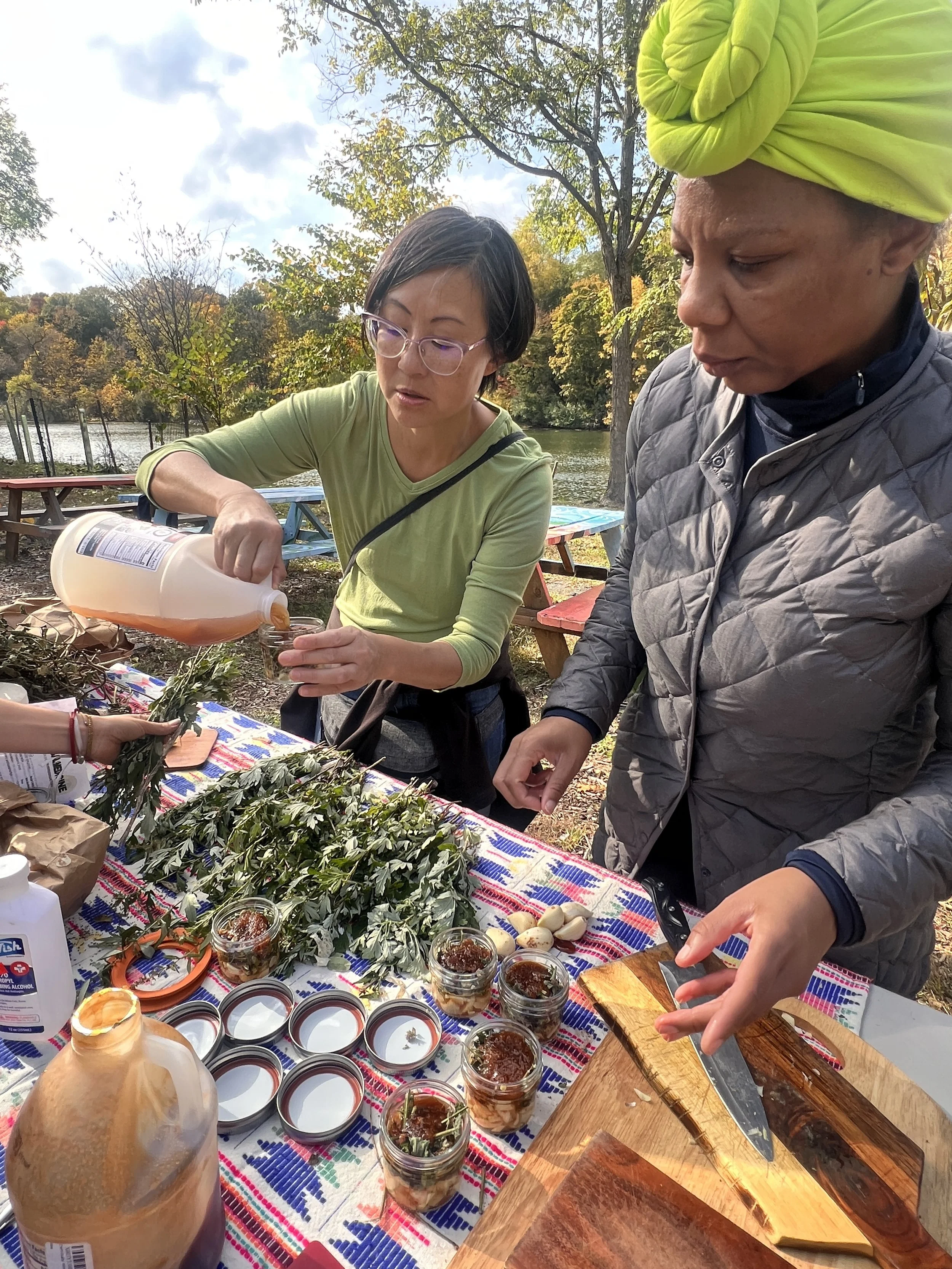 Two women preparing food outdoors on a colorful tablecloth near a body of water and trees with fall foliage. One woman is pouring liquid into small jars, and the other is using a knife on a wooden cutting board. Various jars, herbs, garlic, and cooking supplies are on the table.
