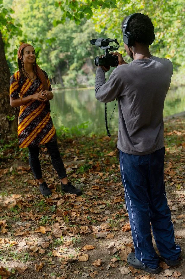 A woman being filmed outdoors by a person with a professional camera near a river surrounded by trees and fallen leaves.