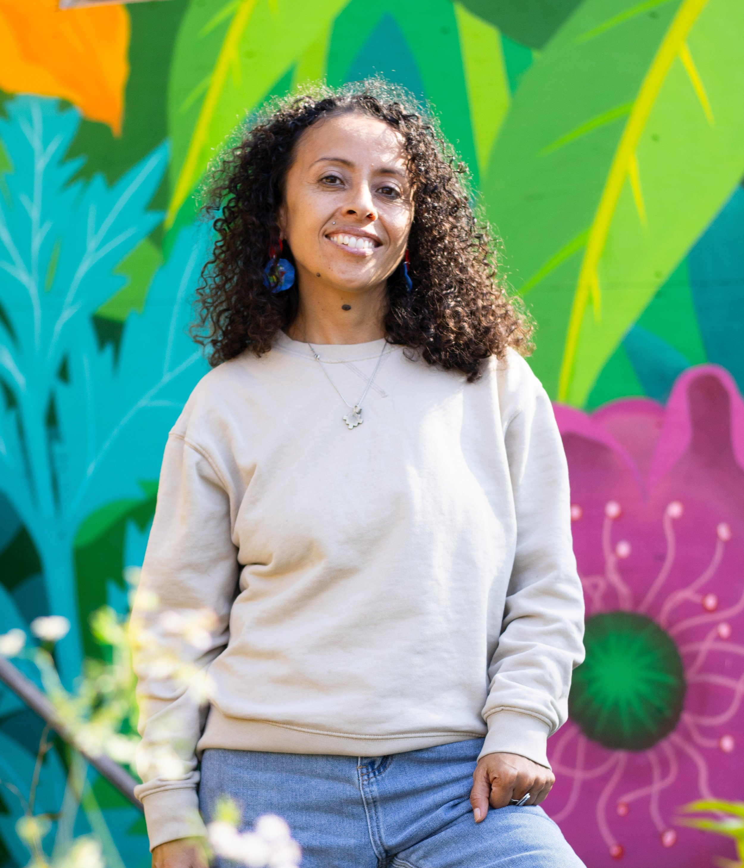 A woman with curly dark hair smiling, wearing a beige sweatshirt and jeans, standing in front of a colorful mural of abstract flowers and leaves.