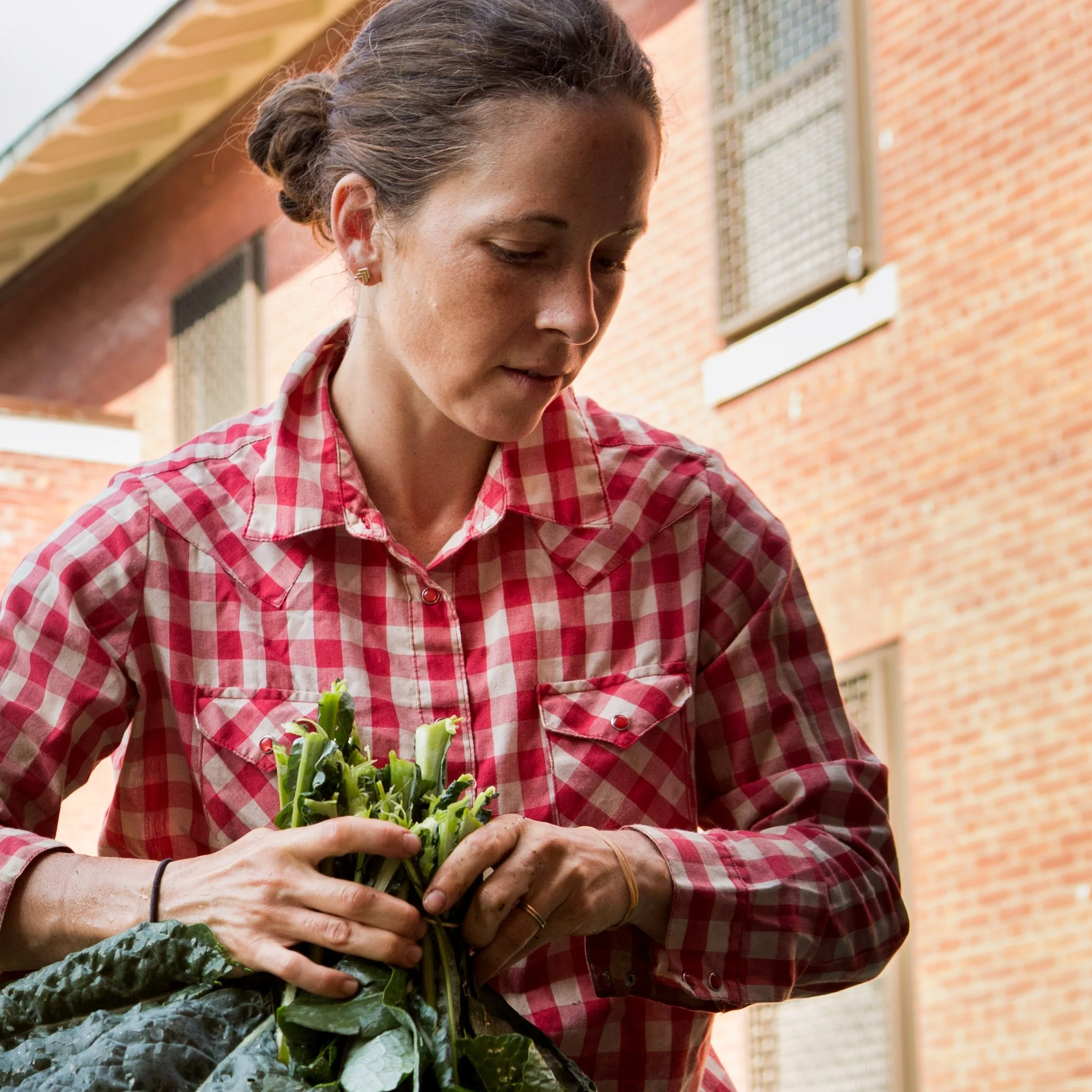 A woman in a red and white checkered shirt holds freshly harvested green vegetables outdoors in front of a brick building.