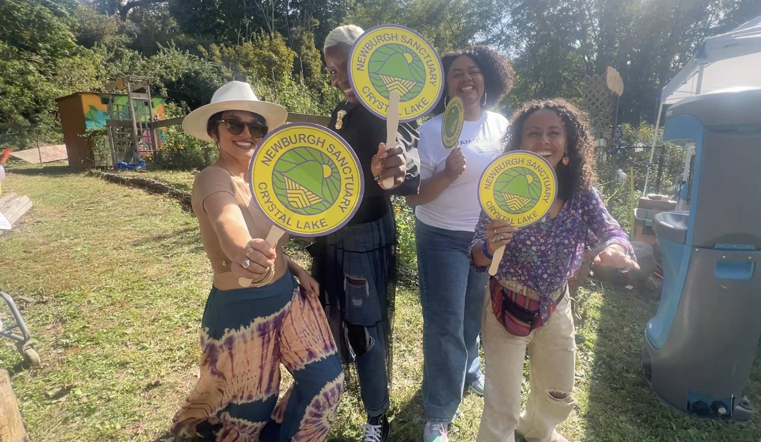 Four women smiling outdoors holding signs with the text 'Newburgh Sanctuary, Crystal Lake' during a sunny day.