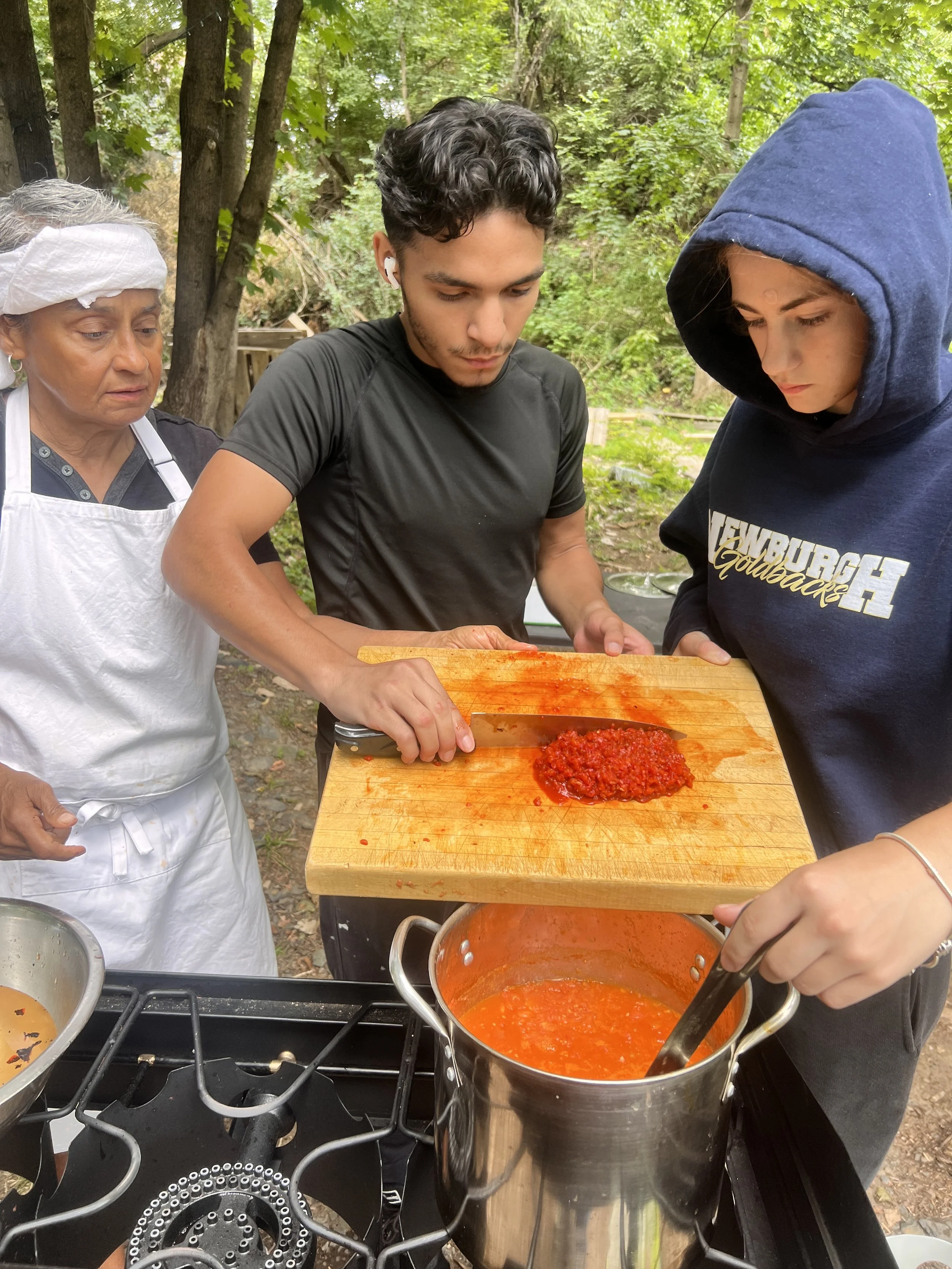 Three people outdoors in a wooded area preparing food. One woman on the left, wearing a white apron and head covering, observes. A young man in the middle, wearing a black athletic shirt, is chopping red sauce on a wooden cutting board. A young woman on the right, dressed in a navy hoodie, is stirring a pot of red sauce on a portable stove.
