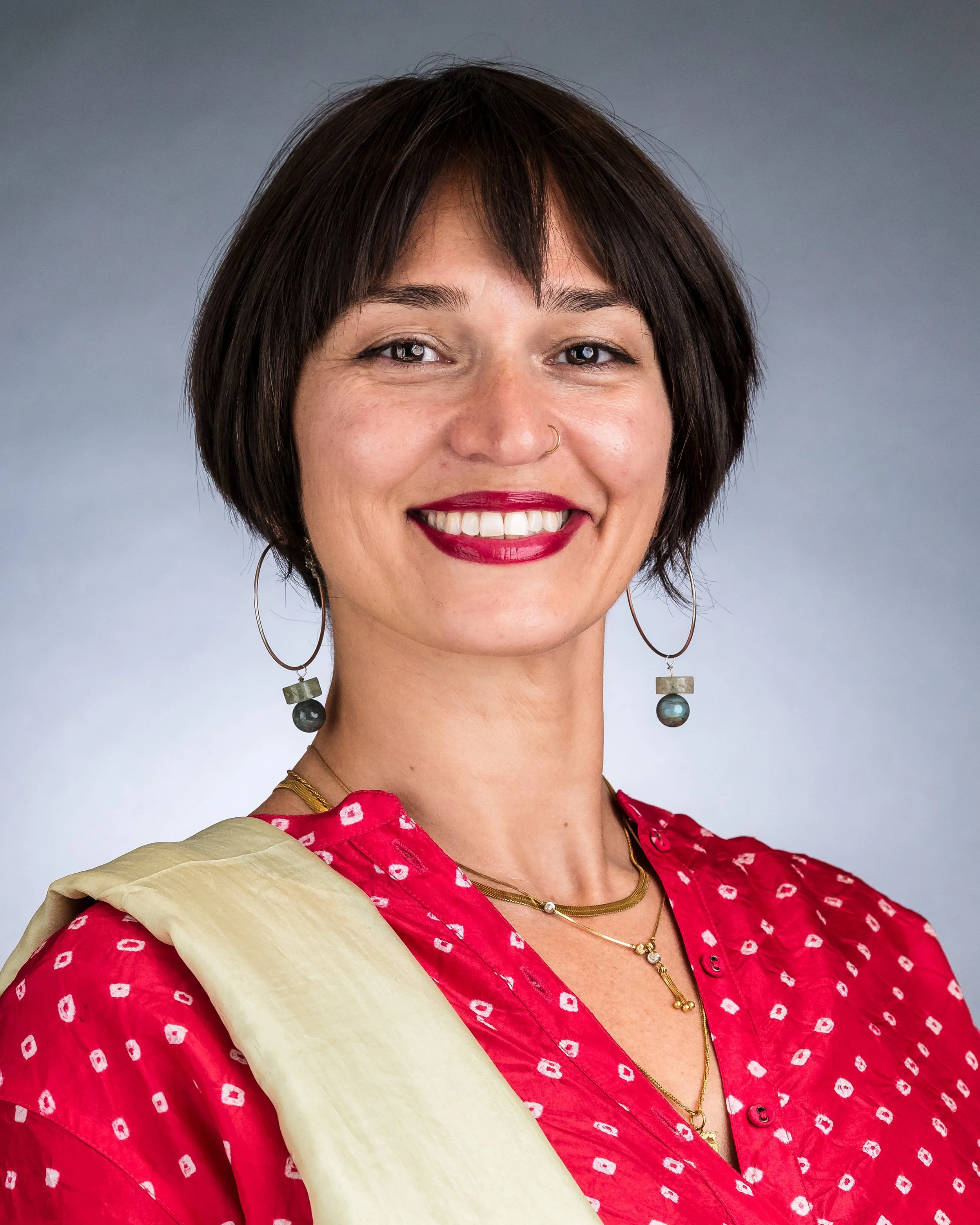 A woman with short dark hair, wearing a red shirt with a white pattern, gold jewelry, and earrings, smiling against a neutral background.