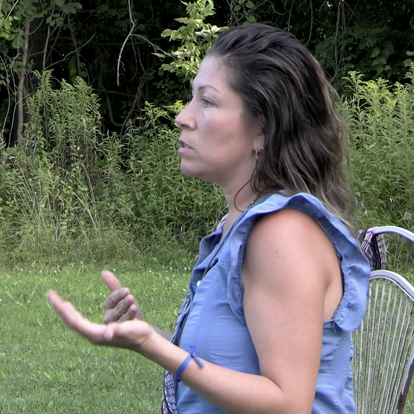 A woman with dark, wavy hair and a nose piercing, wearing a sleeveless blue top, sitting outdoors on a metal chair with greenery in the background, speaking or explaining something.