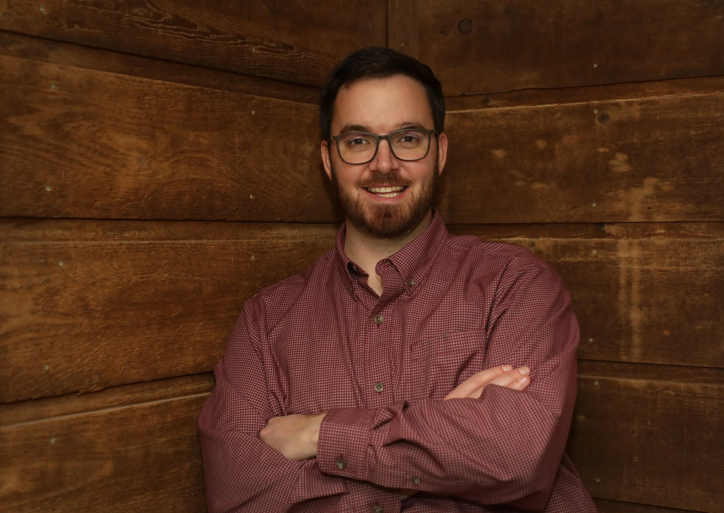 A man with glasses and a beard smiling with arms crossed, standing in front of wooden walls.