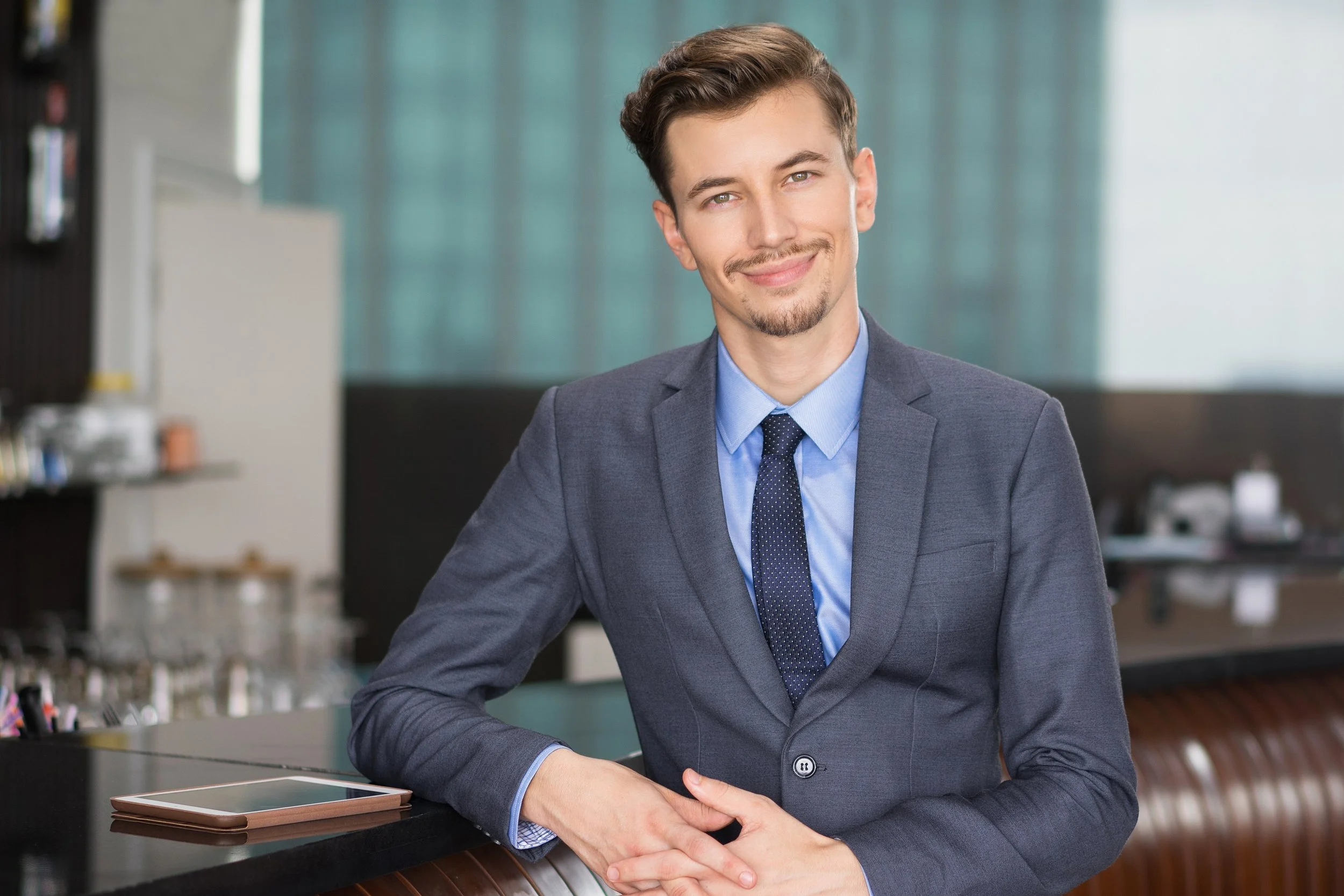 Smiling man in a gray suit leaning on a counter in an office or hotel lobby