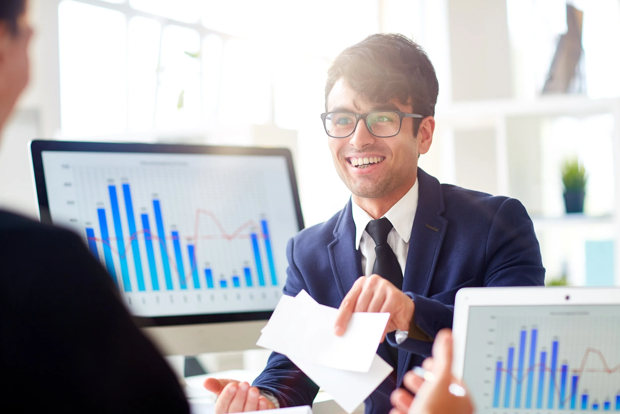 Business professional smiling and speaking during a meeting, with a computer screen displaying bar graphs and charts in the background.