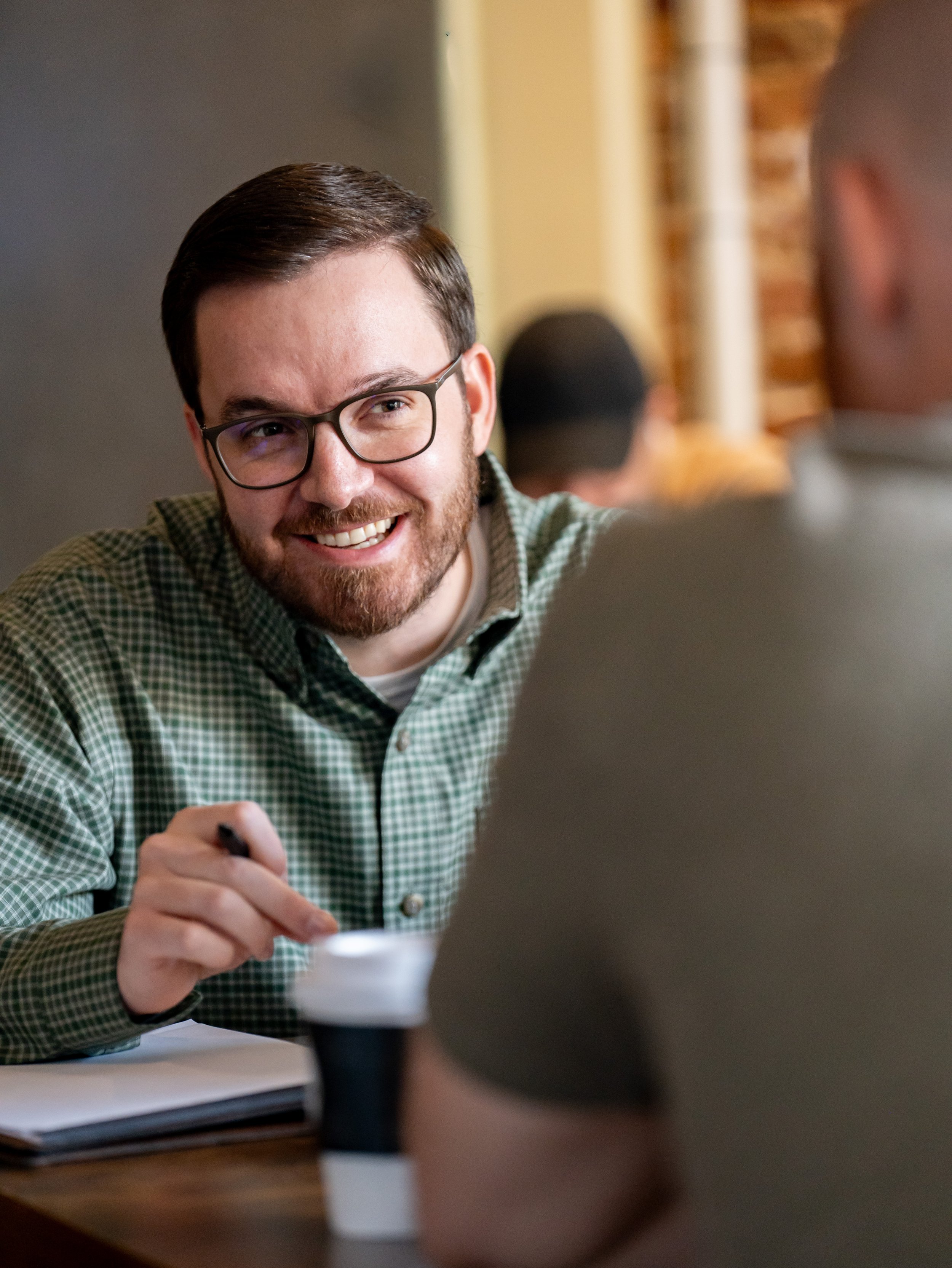 A man with glasses and a beard, smiling and holding a pen, engaging in a conversation with another person at a table in a cafe or restaurant.