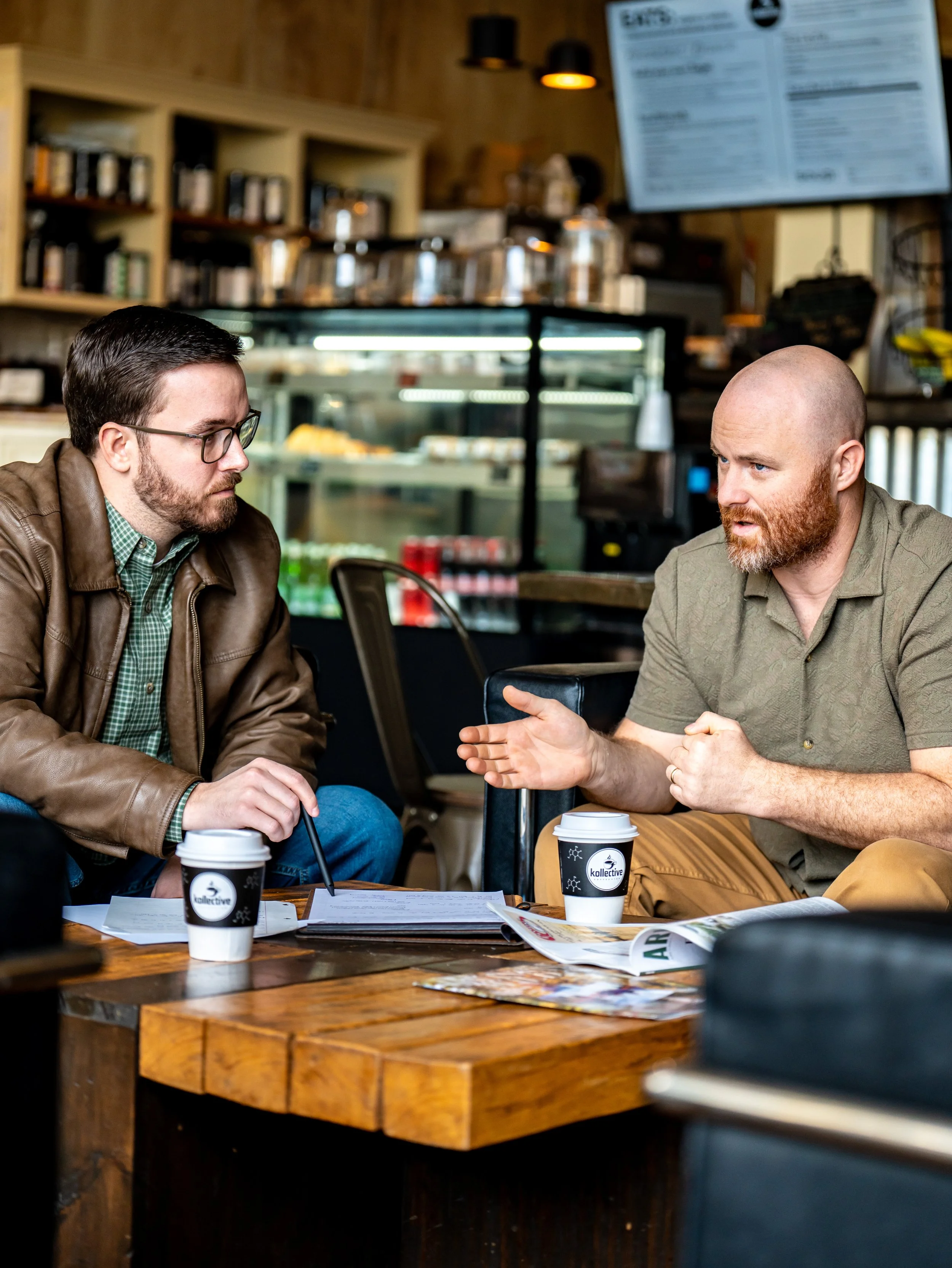 Two men are having a conversation at a wooden table in a coffee shop, with coffee cups, papers, and a tablet on the table, behind a counter with baked goods and a menu board.