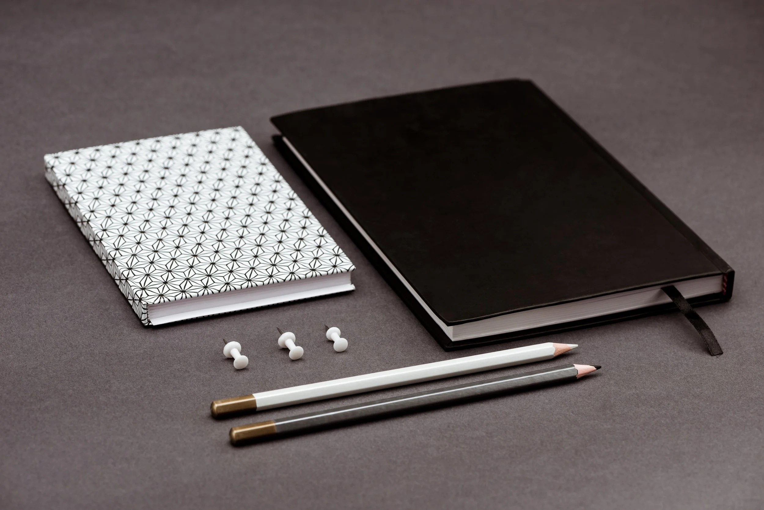 A flat lay of two notebooks, three push pins, and two pens on a gray background.