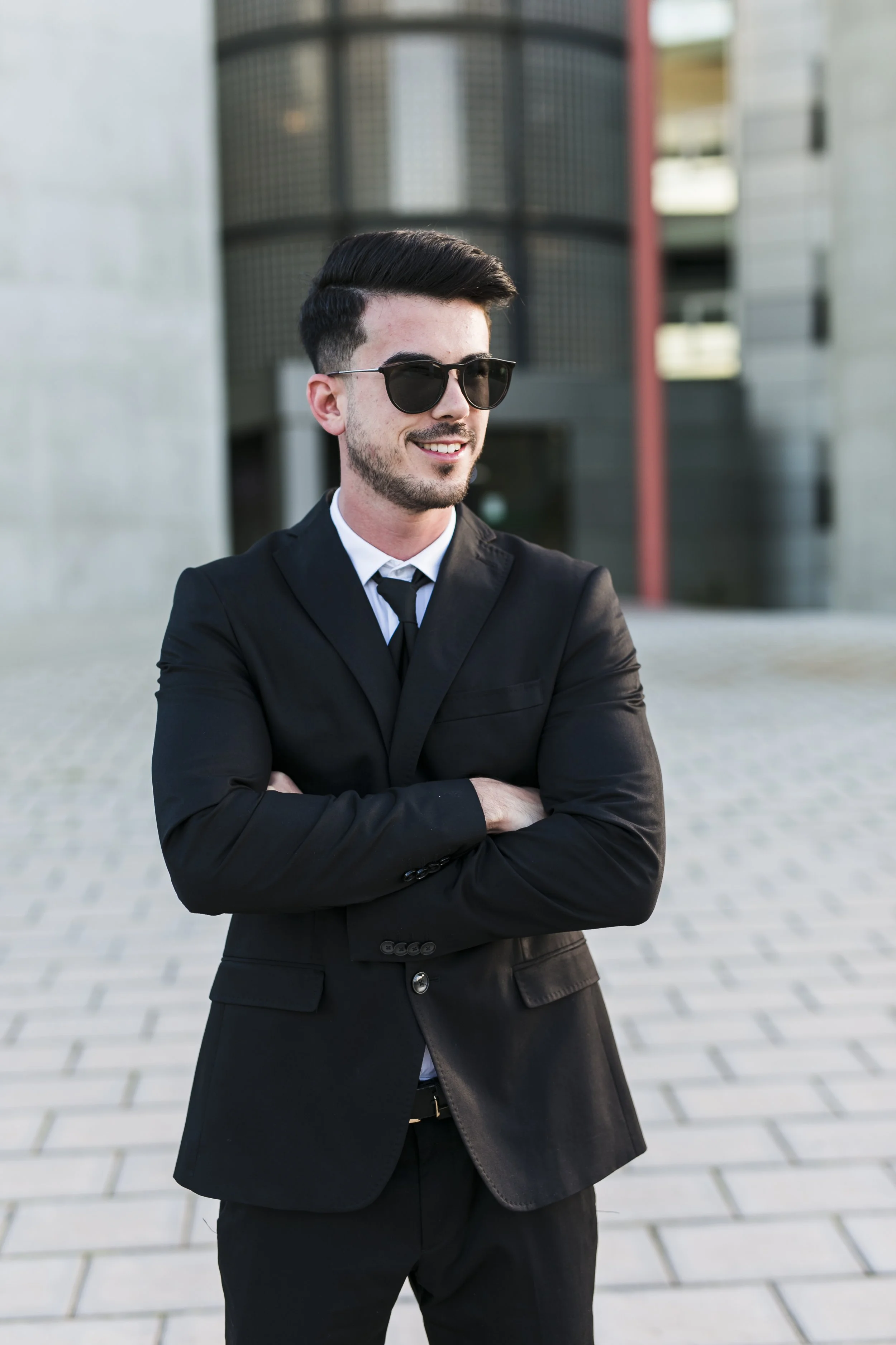 A man in a black suit, white shirt, and black tie standing outdoors with arms crossed, wearing black sunglasses, smiling against a modern building background.