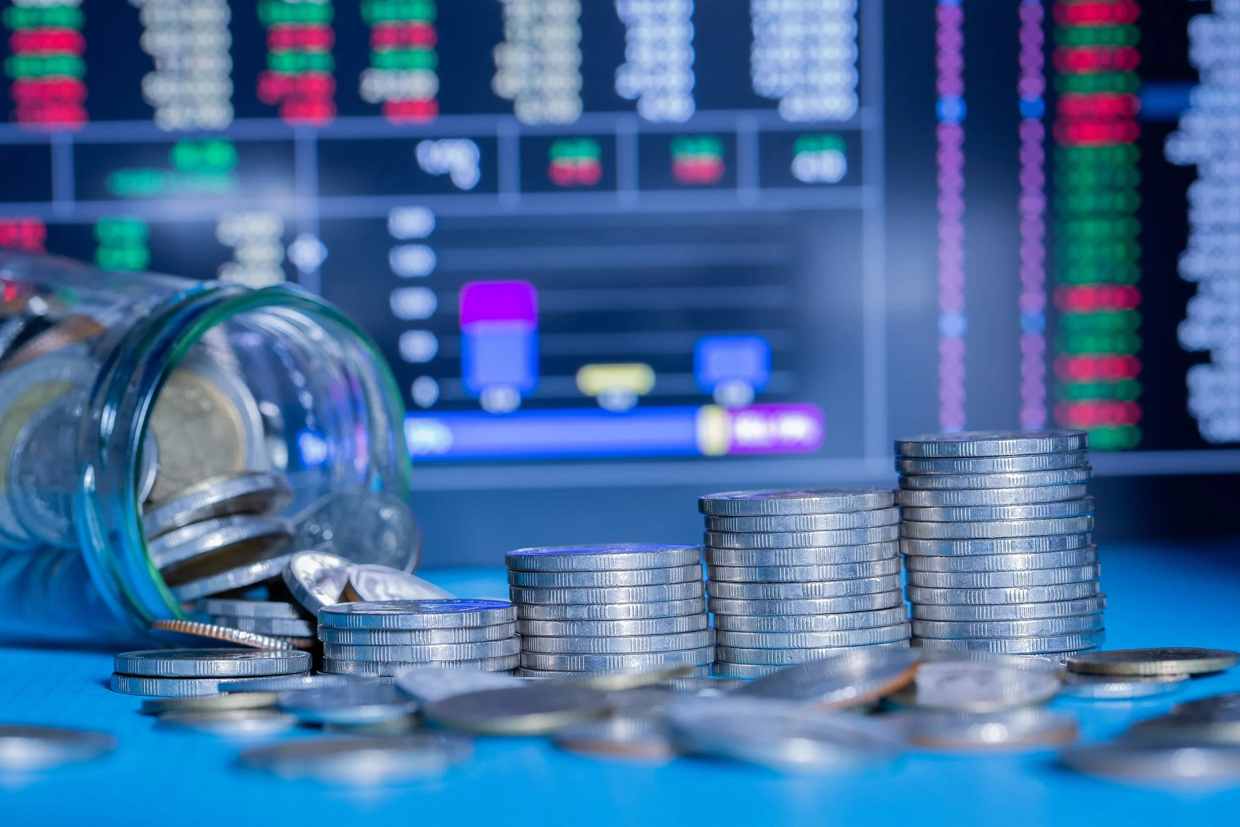 Coins stacked in front of a financial chart on a screen, with some coins spilled from a glass jar.