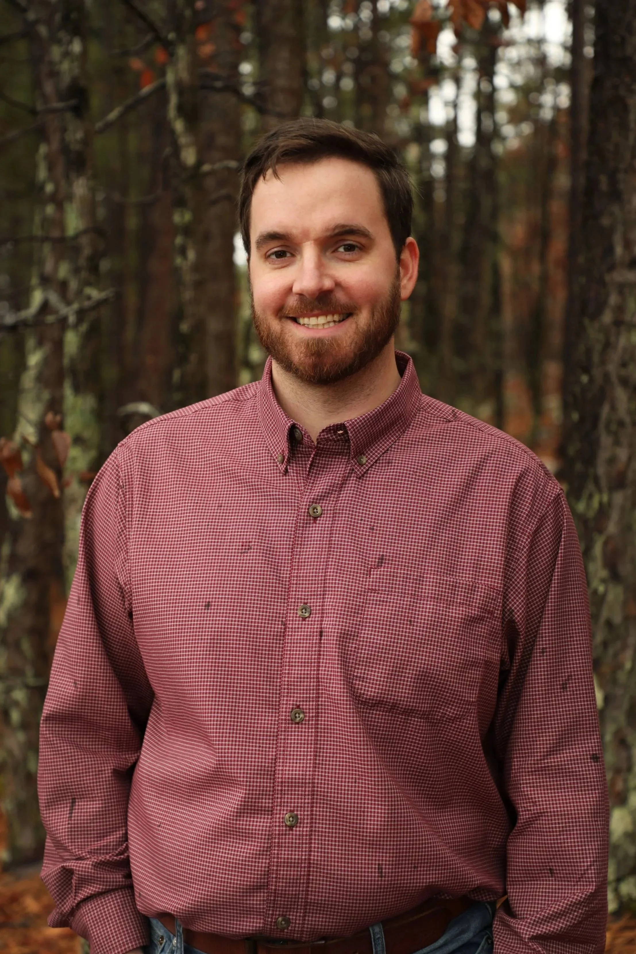 A smiling young man with dark hair and a beard standing outdoors in front of a forested background, wearing a red checkered button-up shirt.