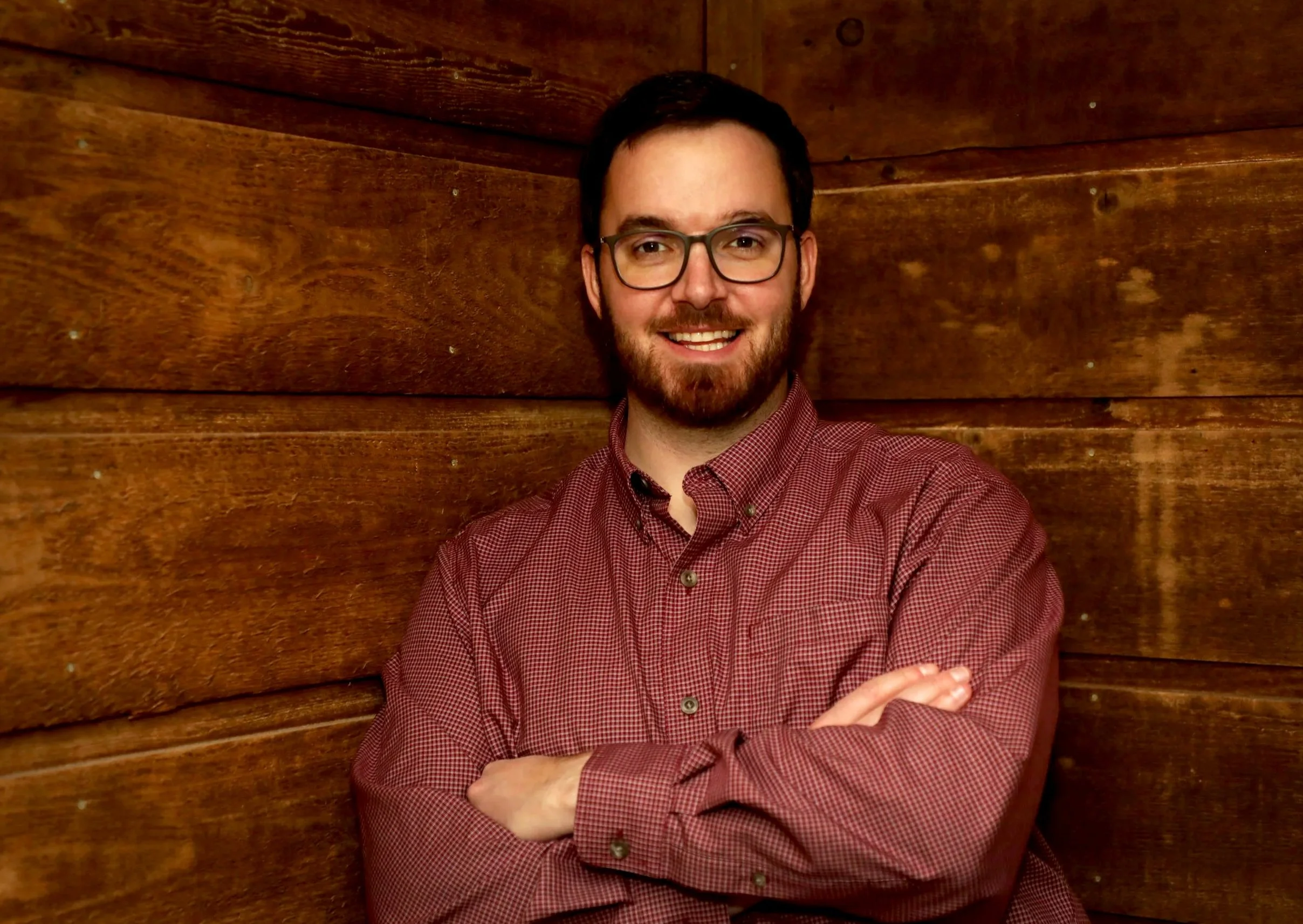 A man with glasses, a beard, and a mustache smiling with arms crossed, standing against a wooden wall.