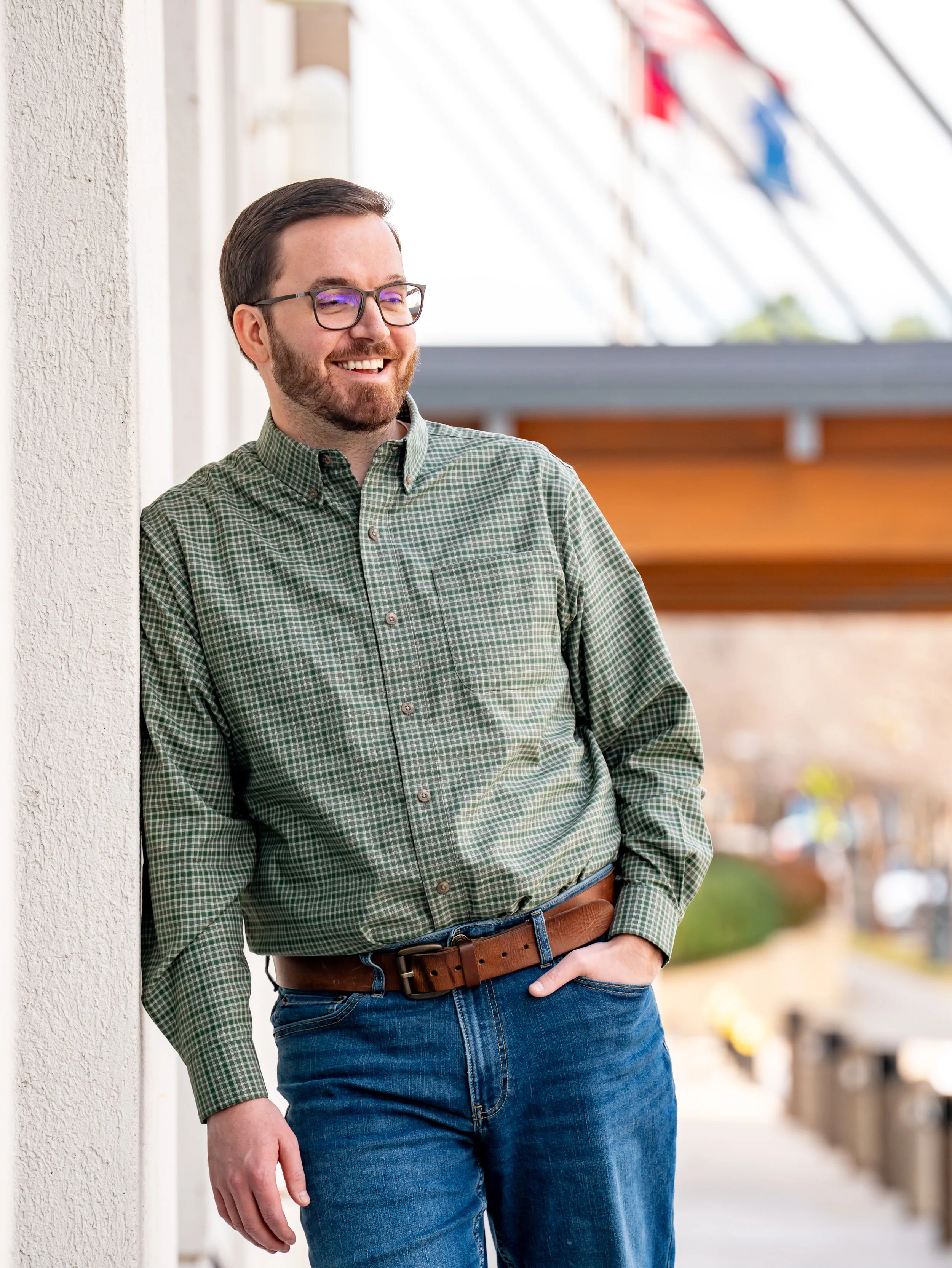 A man with glasses and a beard leaning against a wall outdoors, smiling.