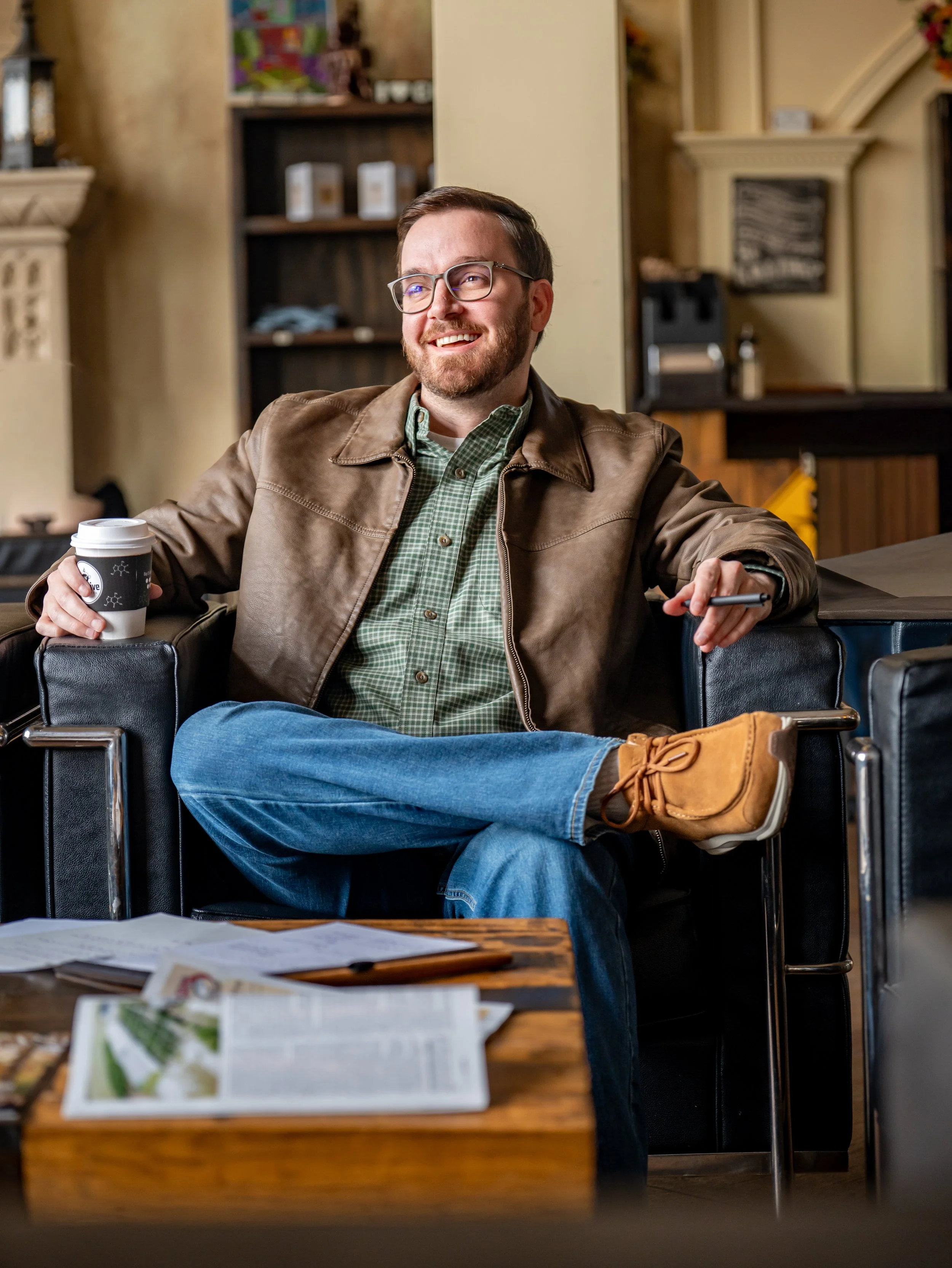 A man sitting in a coffee shop smiling, holding a coffee cup, wearing glasses, a brown leather jacket, green plaid shirt, and tan boots, with papers and magazines on the table in front of him.