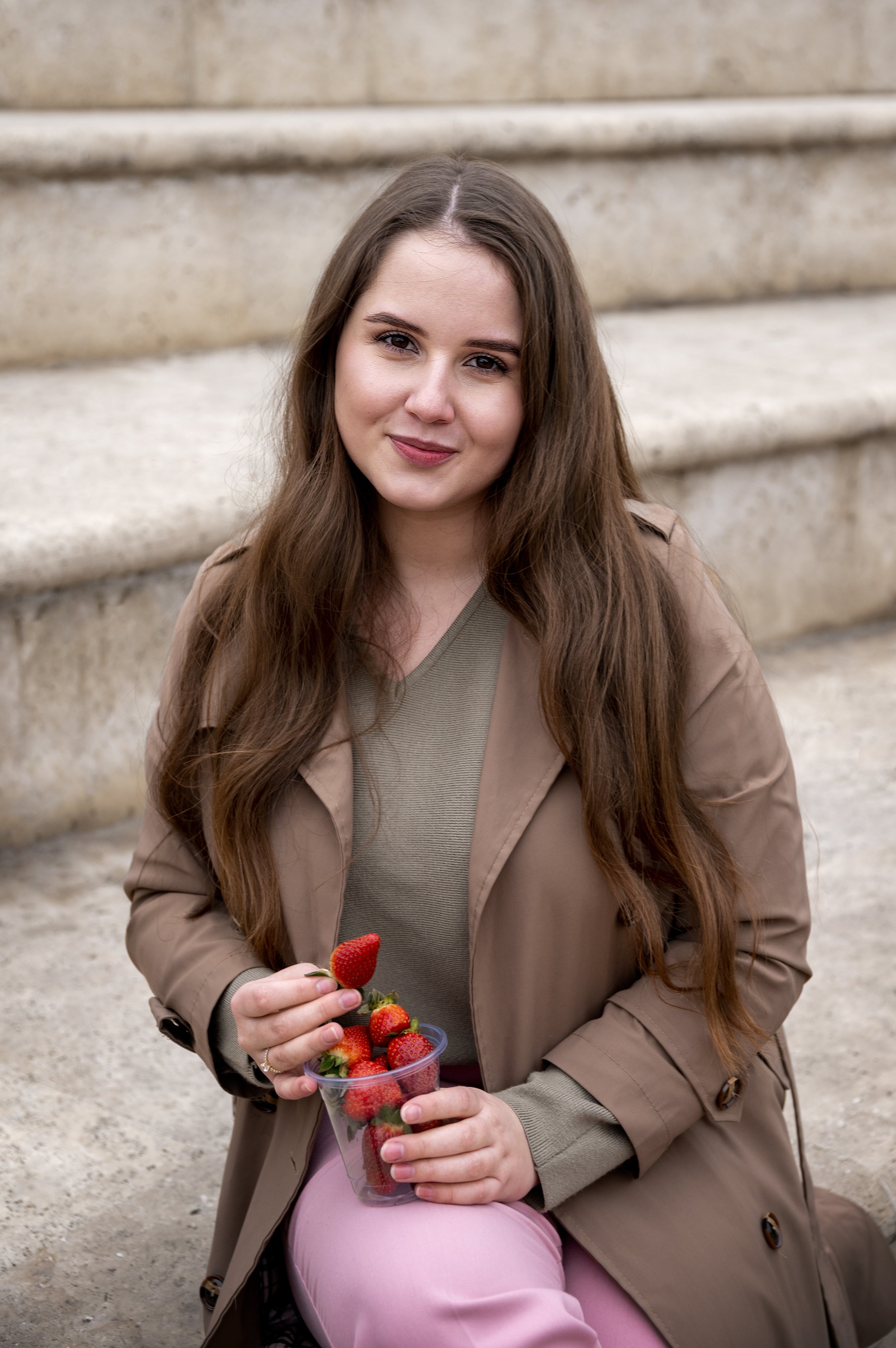 A young woman with long brown hair sitting outdoors on steps, holding a clear cup of strawberries.