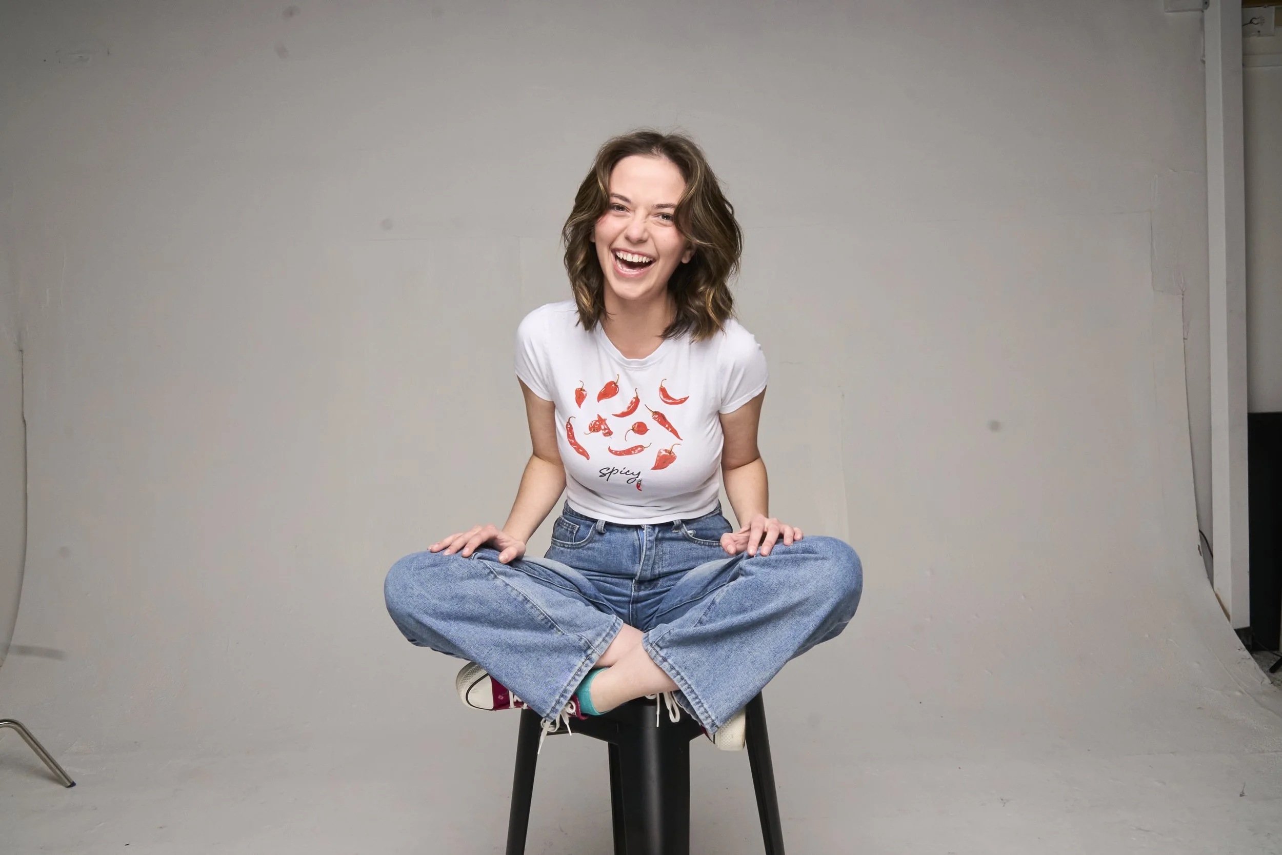 A young woman with shoulder-length brown hair, sitting cross-legged on a black stool, laughing and looking directly at the camera, wearing a white T-shirt with red chili pepper graphics and the word "spicy," and blue jeans, against a plain gray background.