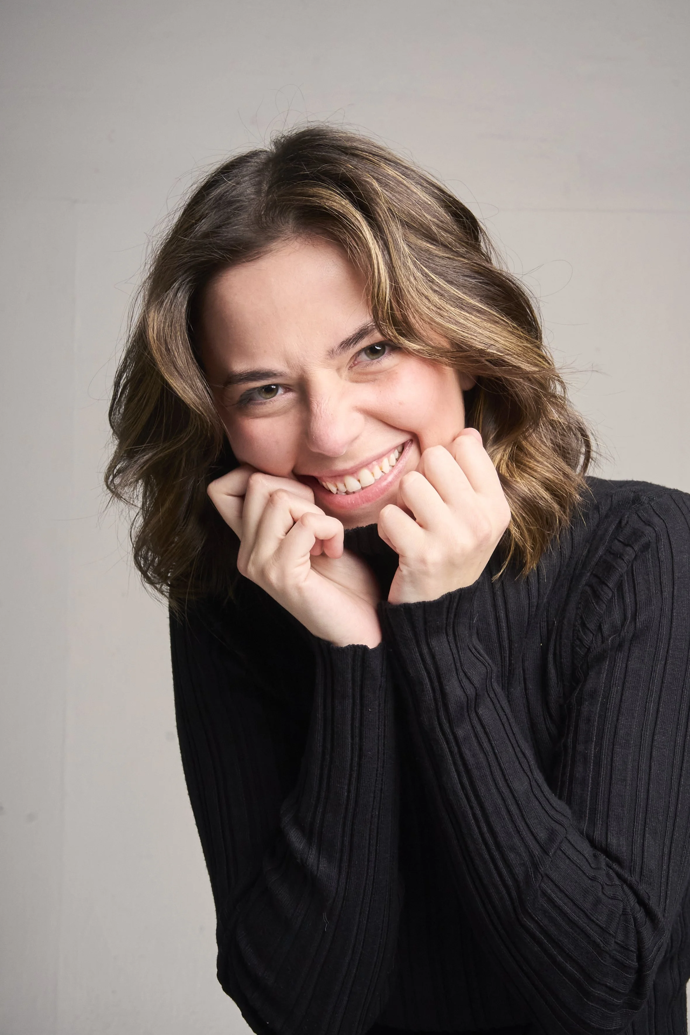 A woman with shoulder-length wavy hair smiling and biting her clenched fists, wearing a black long-sleeve top, against a light gray background.