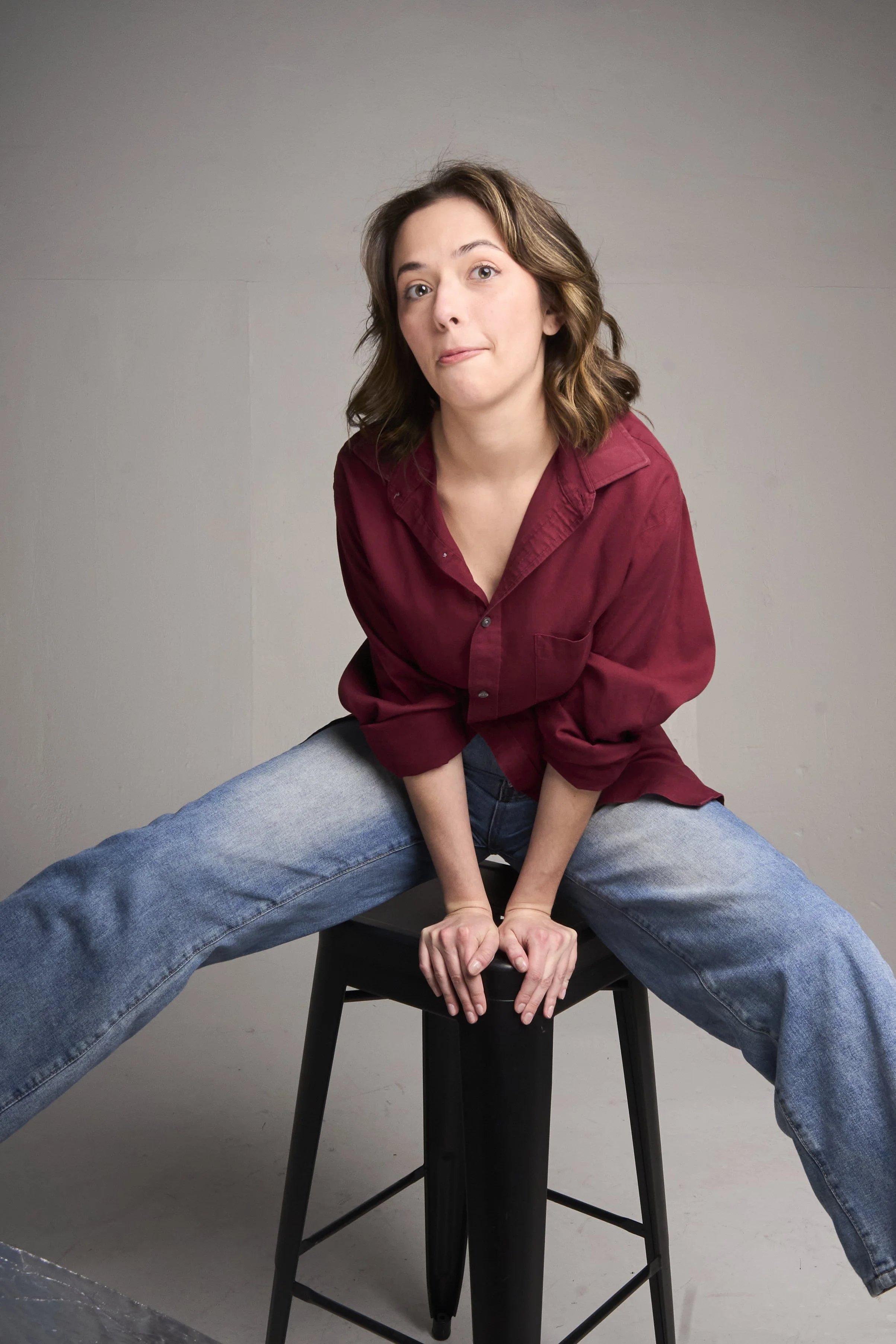 A woman with brown hair wearing a red shirt and blue jeans, sitting on a black stool with legs spread wide, leaning forward with her hands on the stool, posing against a plain gray background.
