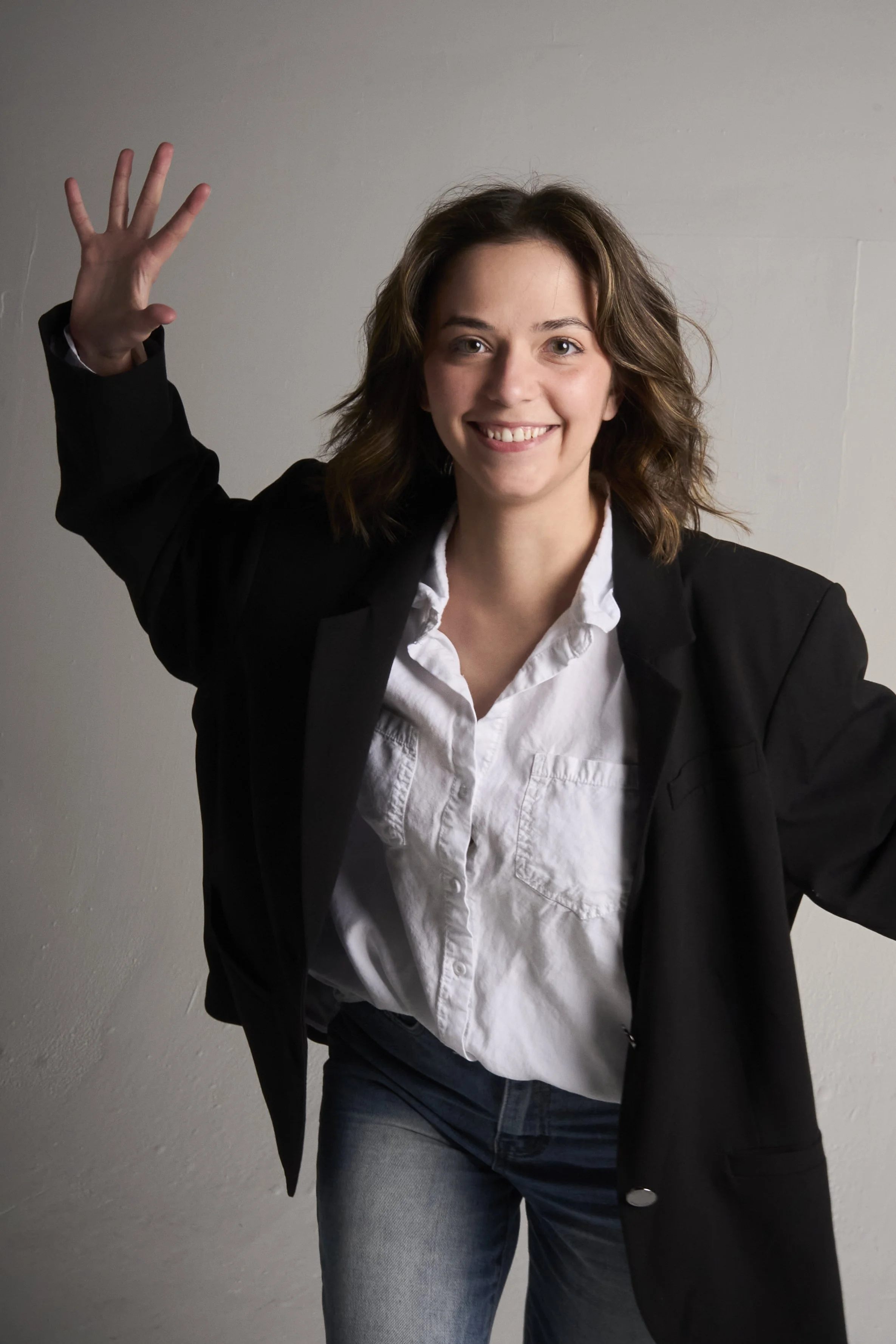 A young woman with shoulder-length brown hair, smiling, wearing a black blazer over a white button-up shirt and jeans, standing against a plain wall with her right hand raised in a gesture.