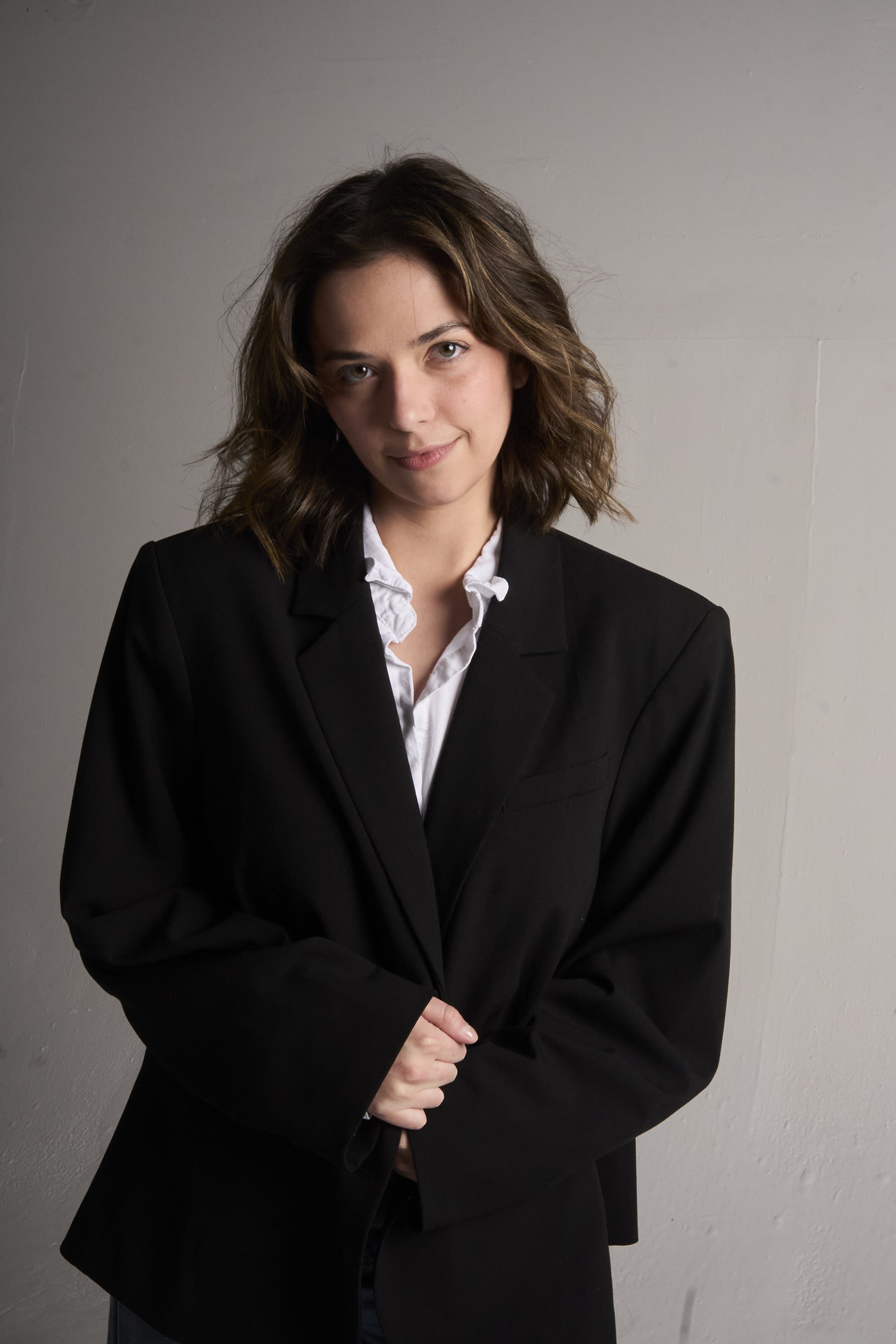 A young woman with shoulder-length wavy brown hair and blue eyes, dressed in a black blazer over a white ruffled shirt, standing against a plain beige wall.