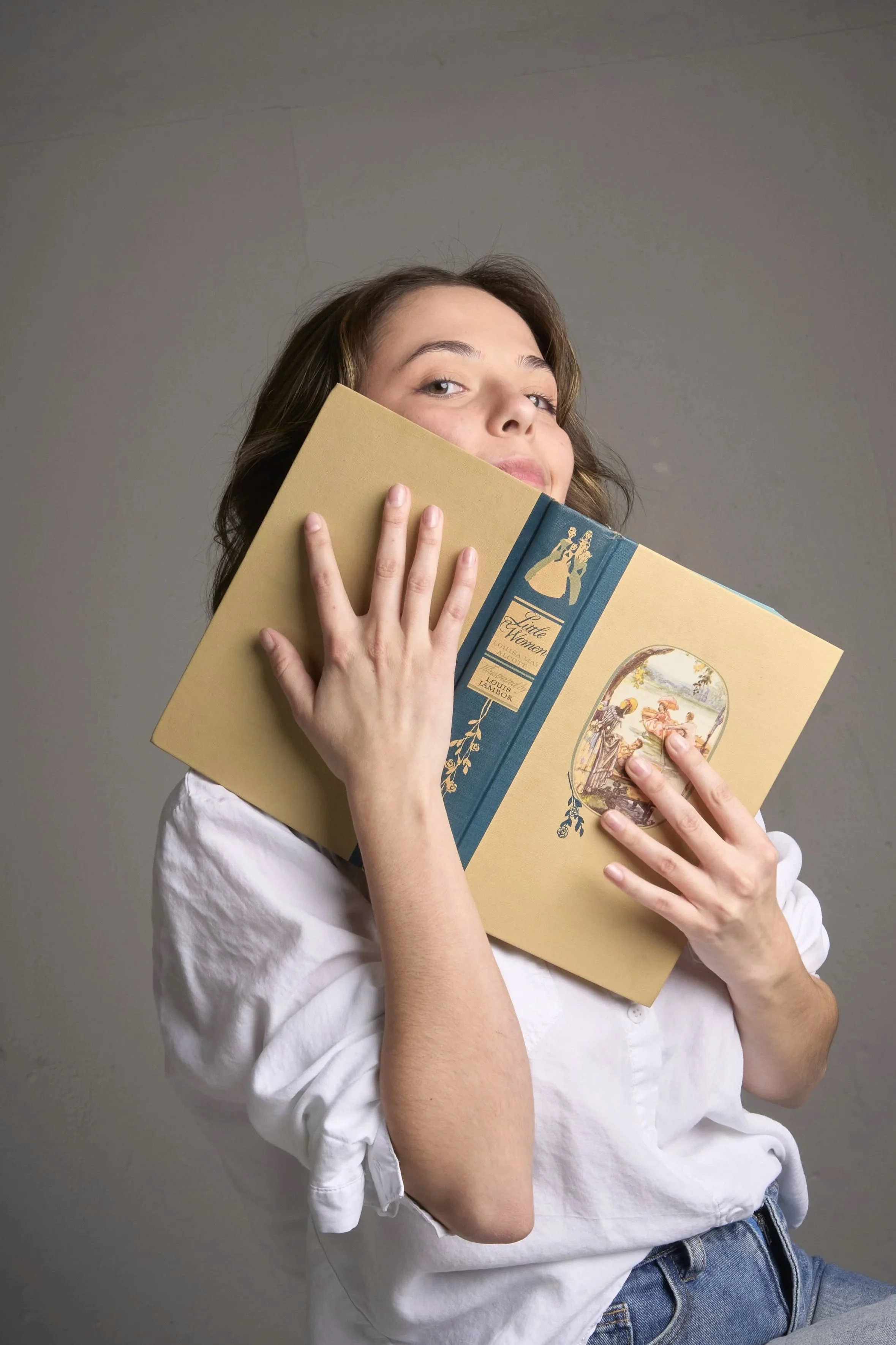 A young woman with brown hair holding a vintage book titled 'Little Women' close to her face, partially covering her cheek.