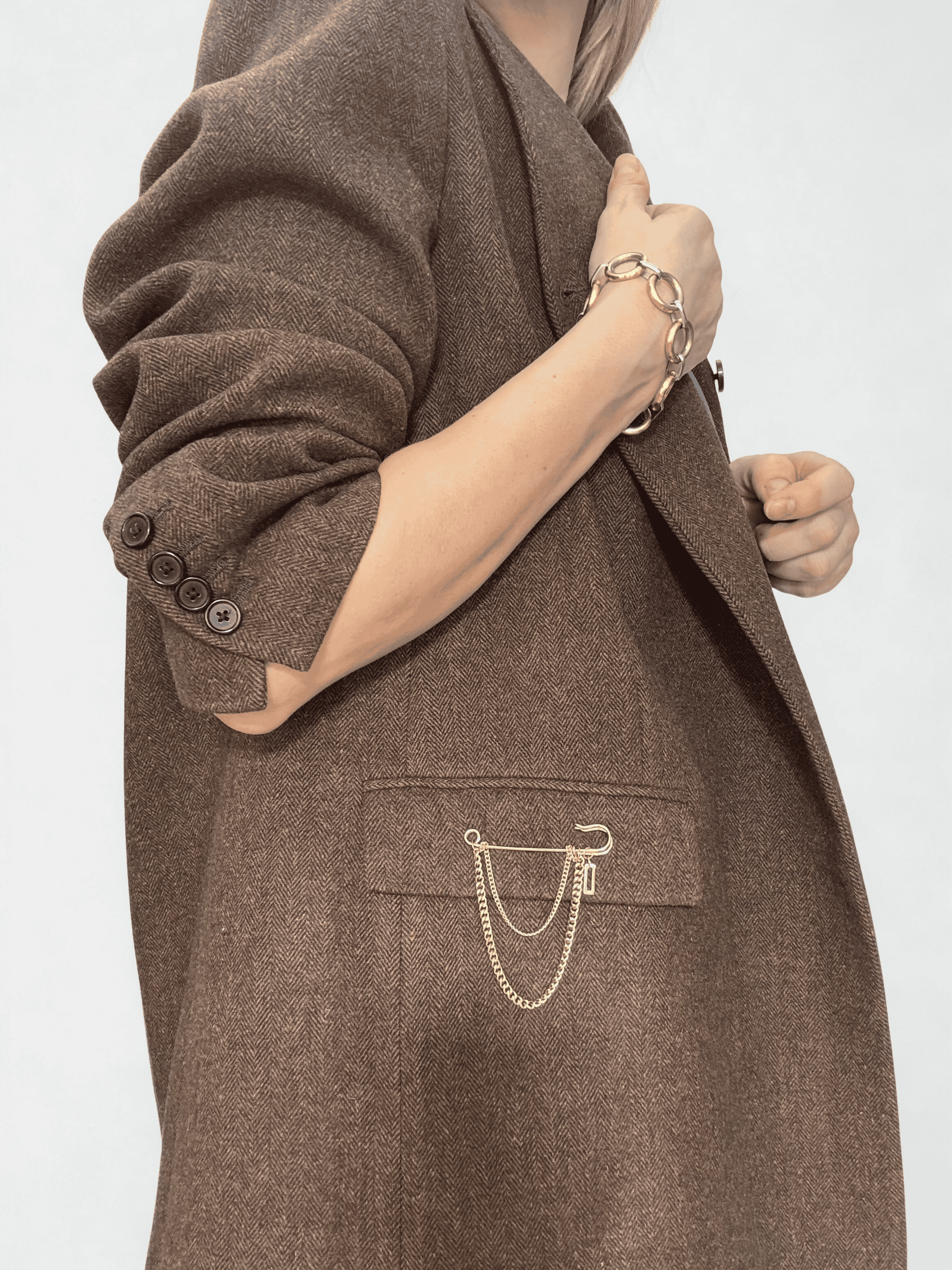 Golden oversized safety pin brooch with a double-tier of delicate curb chains, styled on the pocket of a brown wool blazer.