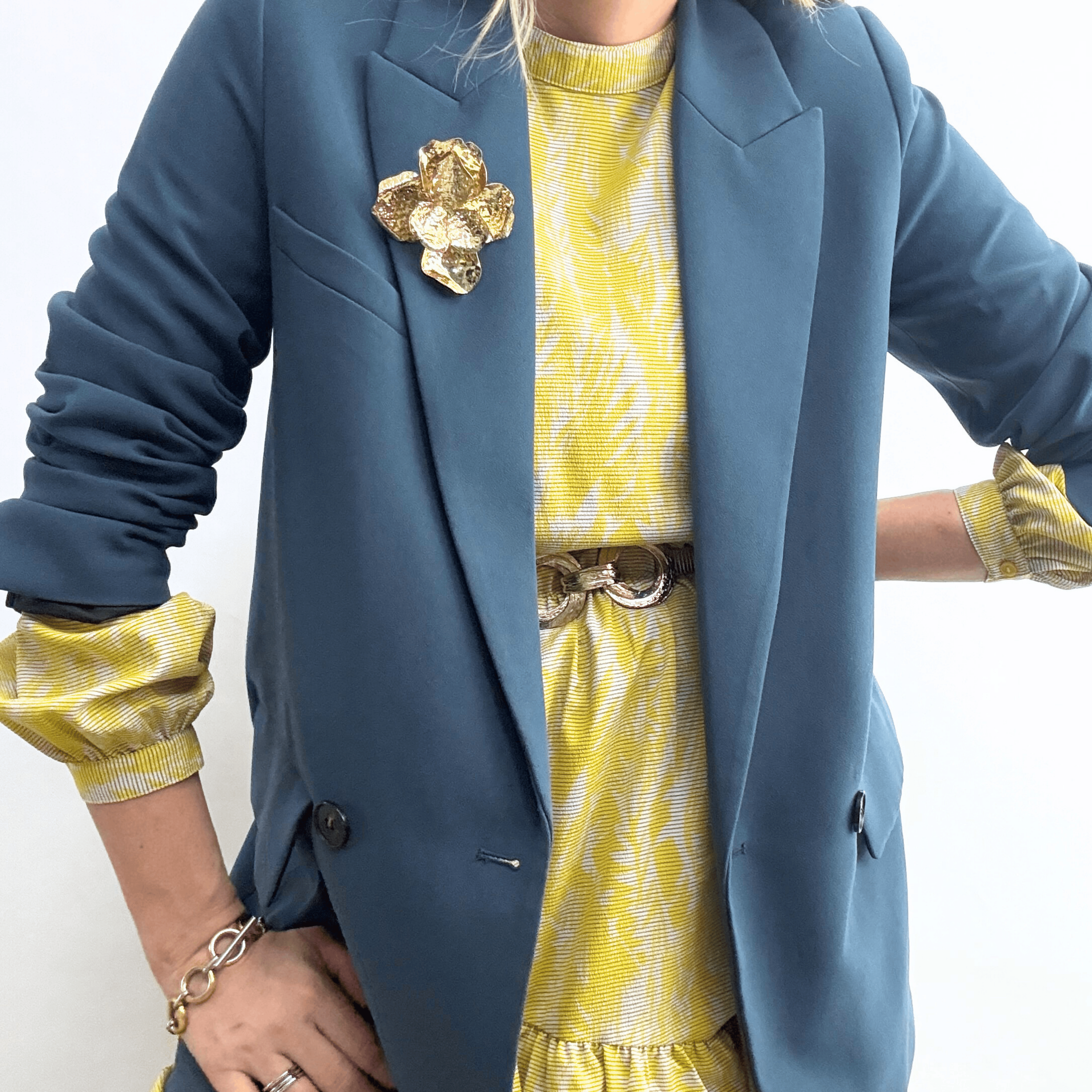 A hammered metal flower brooch in an antique-gold finish styled on a blue blazer with a yellow patterned dress.