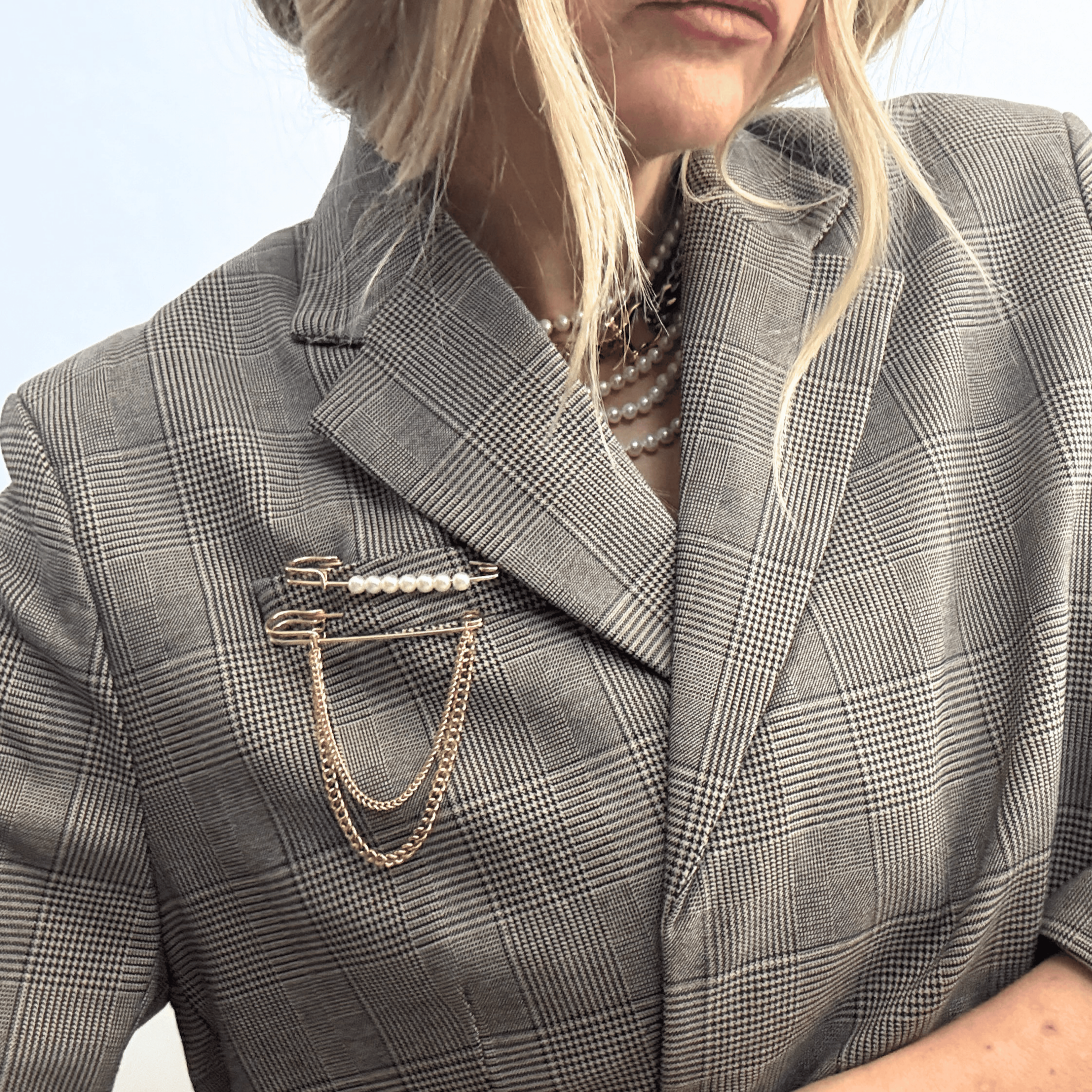 Close-up of a person wearing a grey plaid blazer adorned with pearl and chain brooches, and layered pearl necklaces.