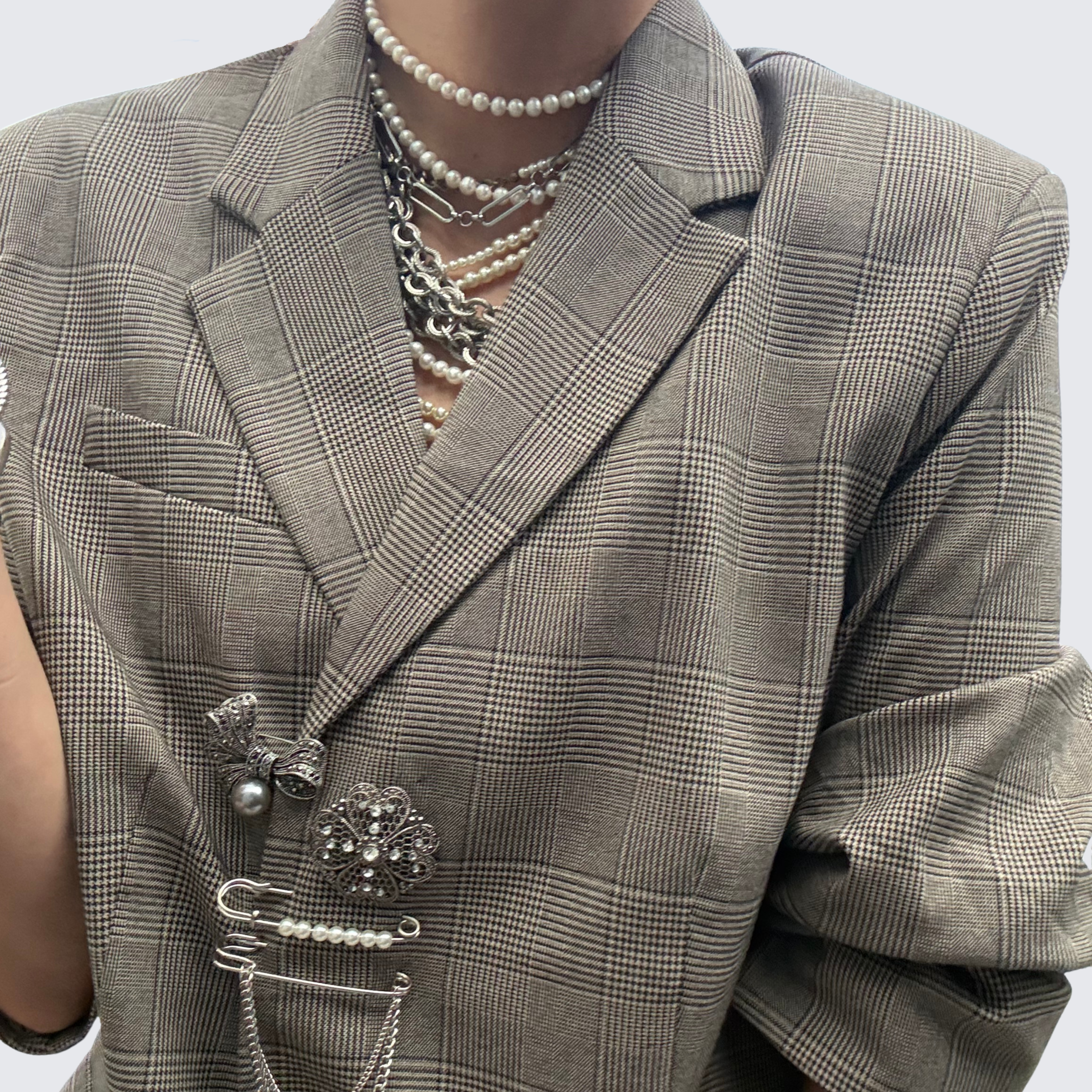 Woman wearing plaid jacked with silver and pearl brooch stack. She also wears a pearl multi strand necklace and chunky silver necklaces.