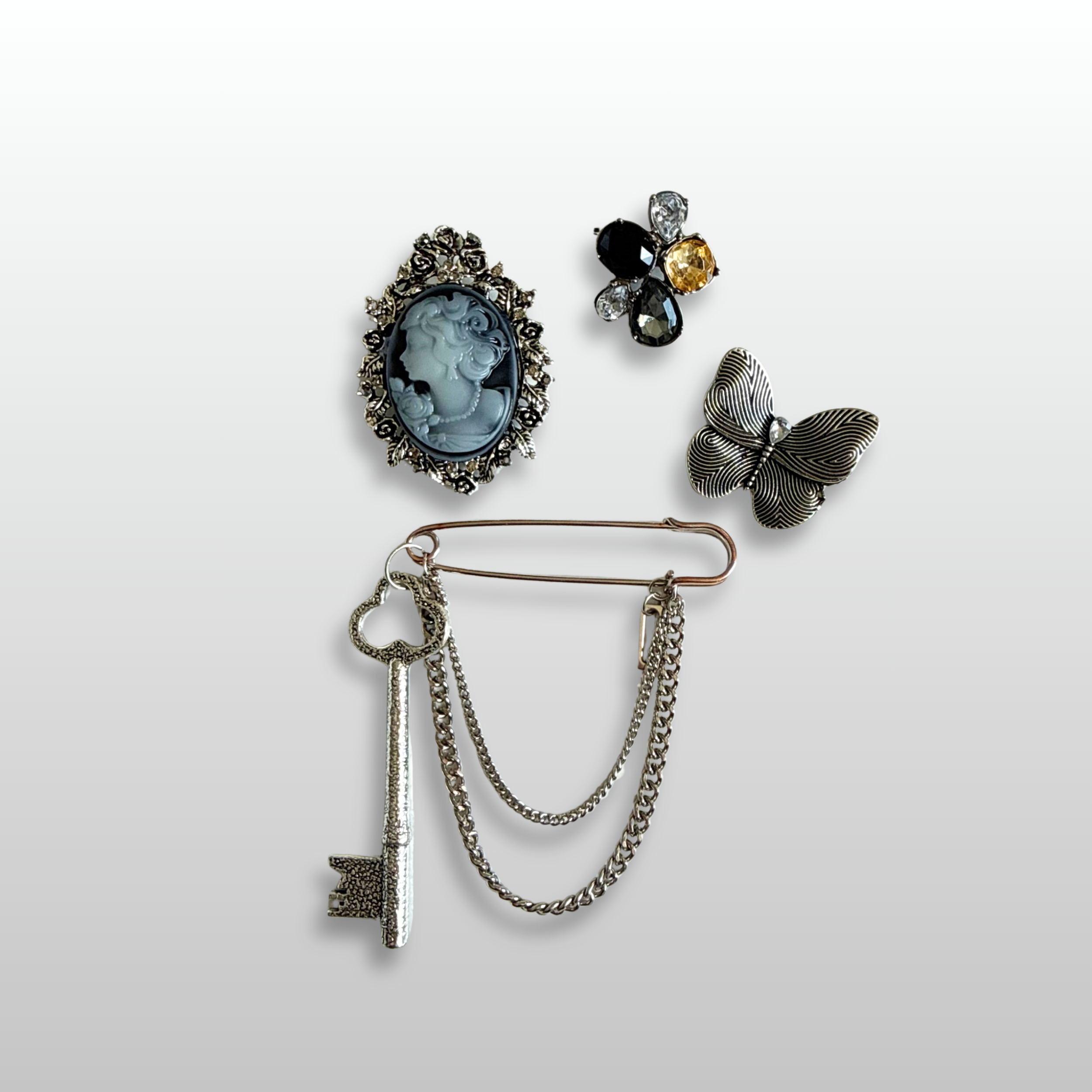 A close up of a The Twillight Heirloom Brooch Stack from The Brooch House. A draped pin with skeleton key charm, a silver butterfly, a small teardrop cluster stud and an indigo and silver tone cameo.
