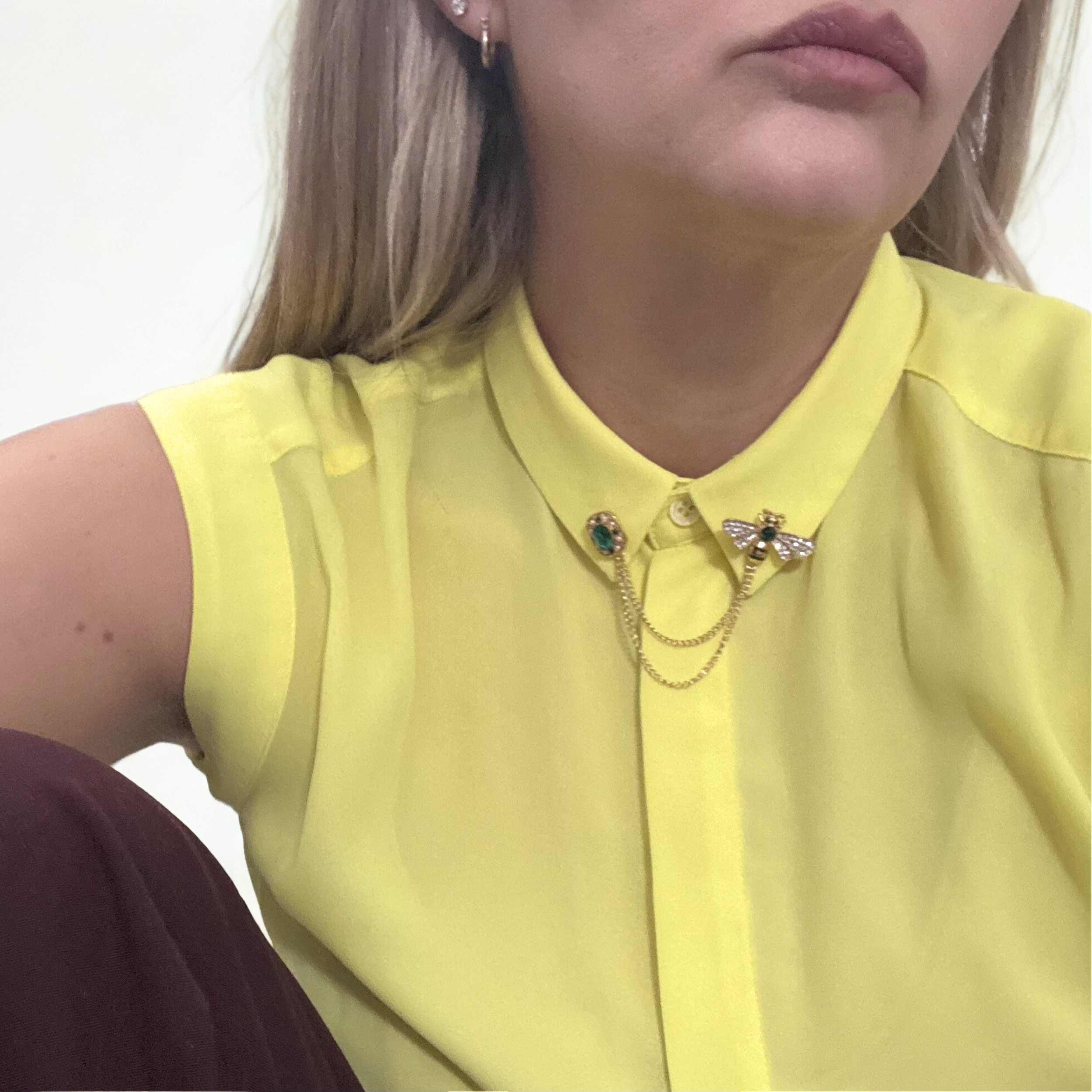 Close-up of a woman wearing a yellow sleeveless blouse with a collar, adorned with a gold chain necklace featuring a bee and an emerald-green gemstone. She also has small hoop earrings and a small stud in one ear.