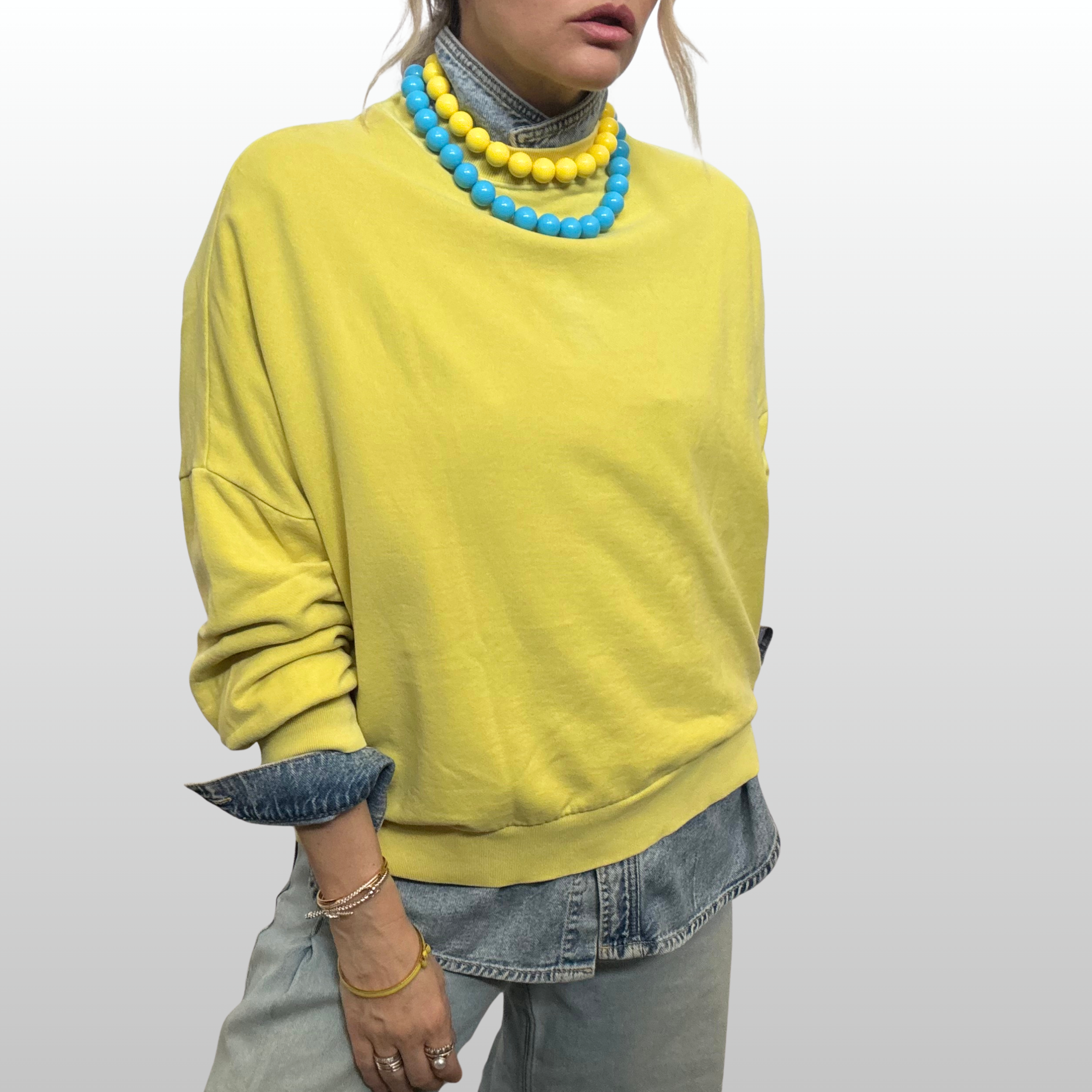 Woman wearing yellow sweatshirt layered over denim shirt and and jeans. She is wearing two strands of resin beads, one blue and one yellow.