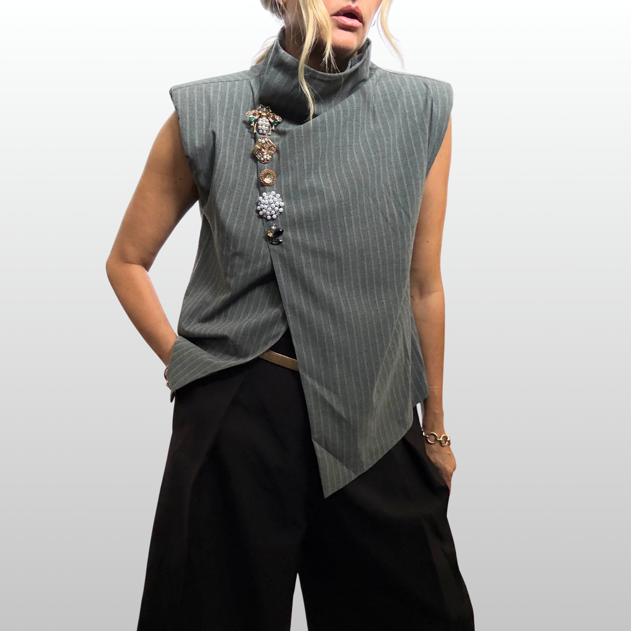 Close up of woman wearing an asymmetric sleeves grey pin striped vest. Ar the closure of the vest is a vertical stack of 5 brooches in various gold and amber tones.